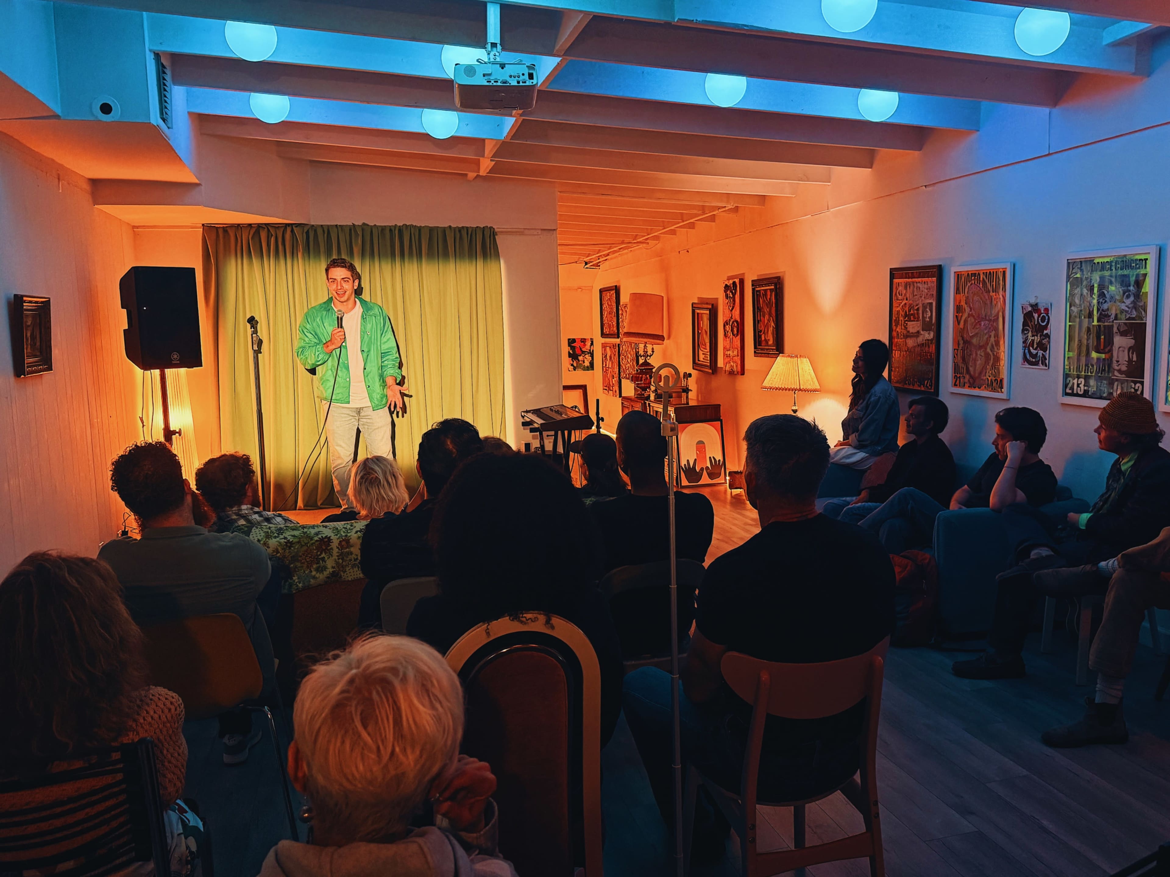 A comedian performs on stage in a well-lit room filled with an audience sitting on chairs and benches, surrounded by colorful artwork on the walls.