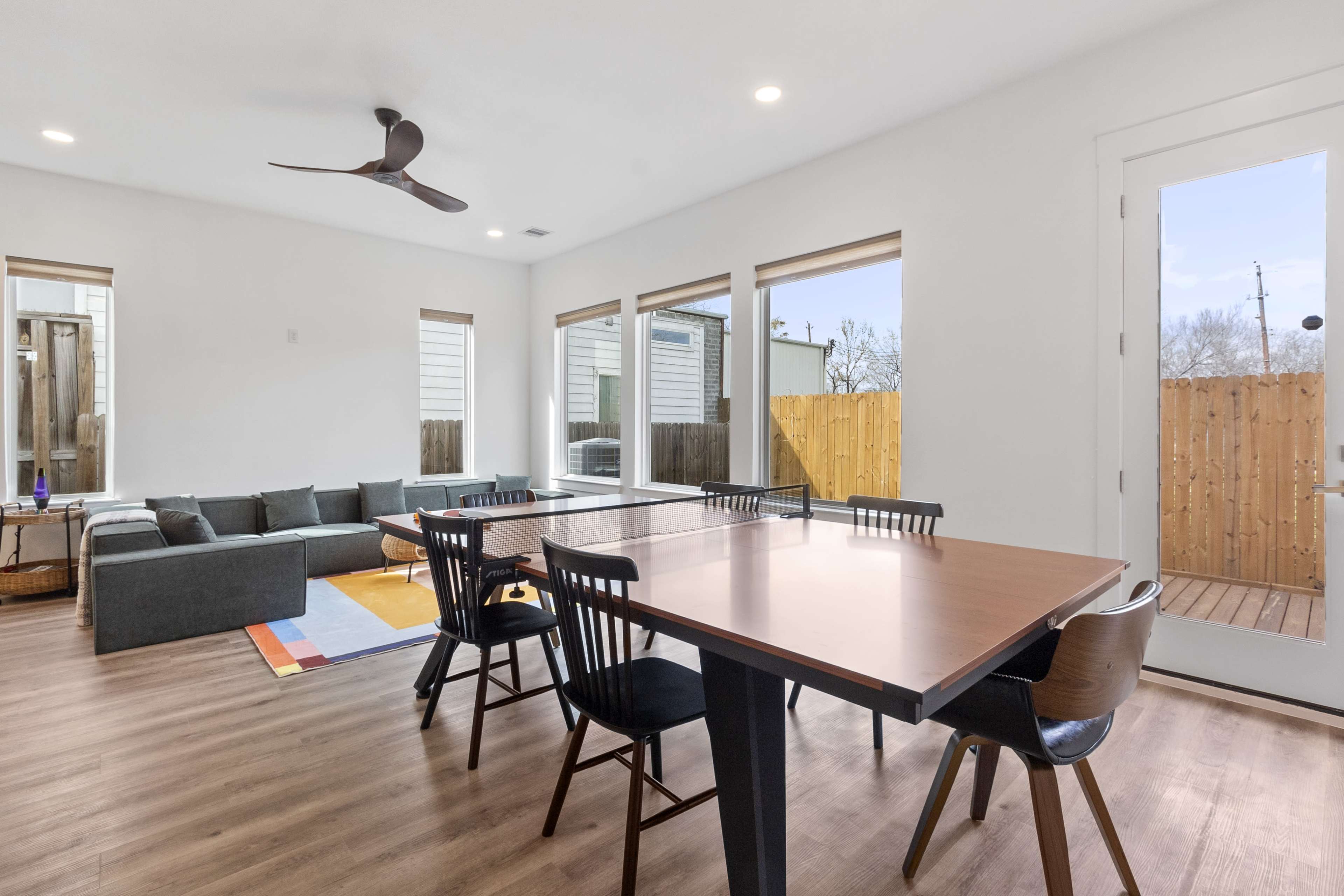 The image shows a spacious, modern indoor living area featuring a dining table, chairs, a sectional sofa, and large windows overlooking a fenced outdoor space.