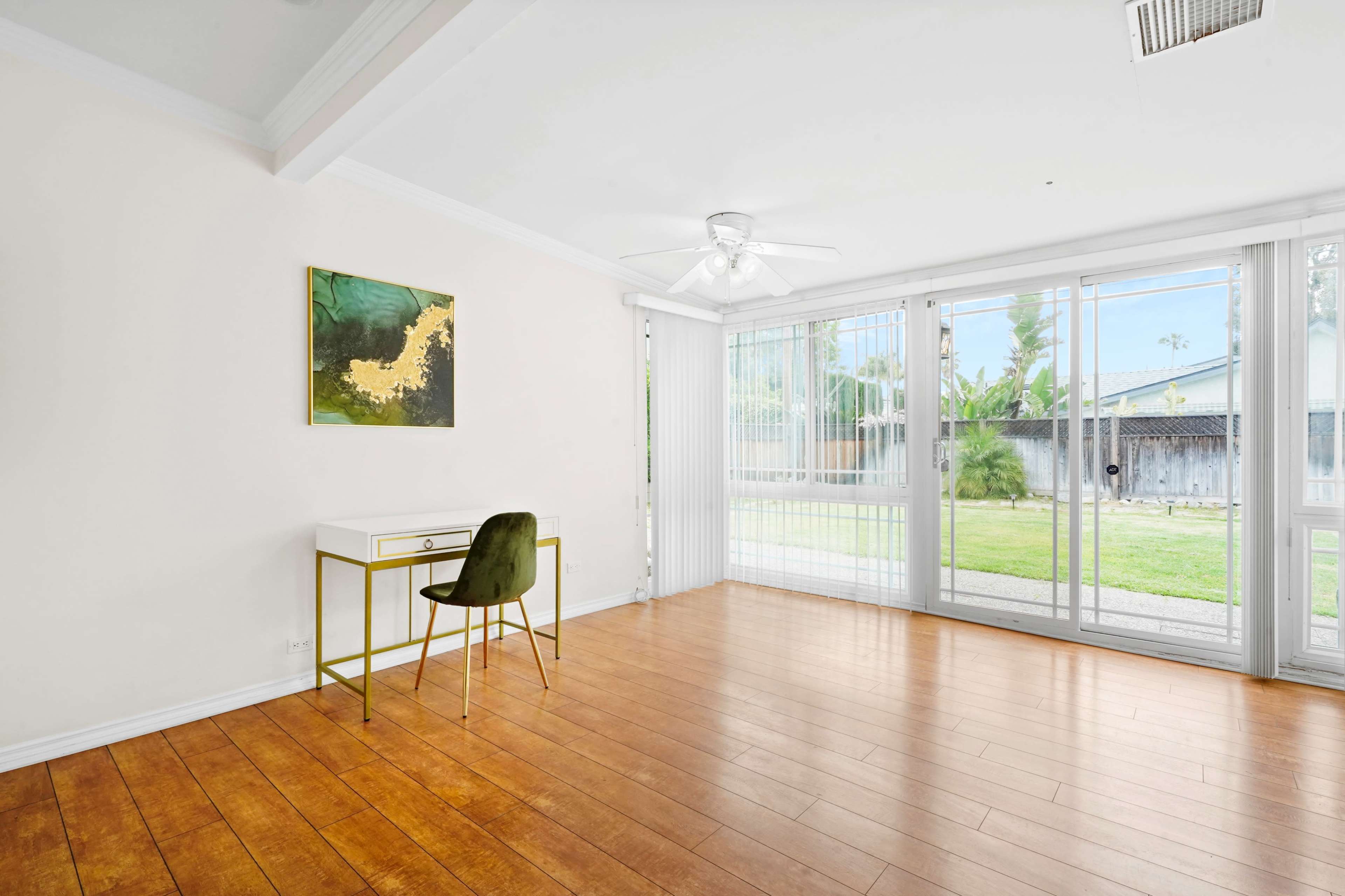 A bright room features a wooden floor, a white desk with a chair, and large sliding glass doors that lead to a grassy outdoor area.