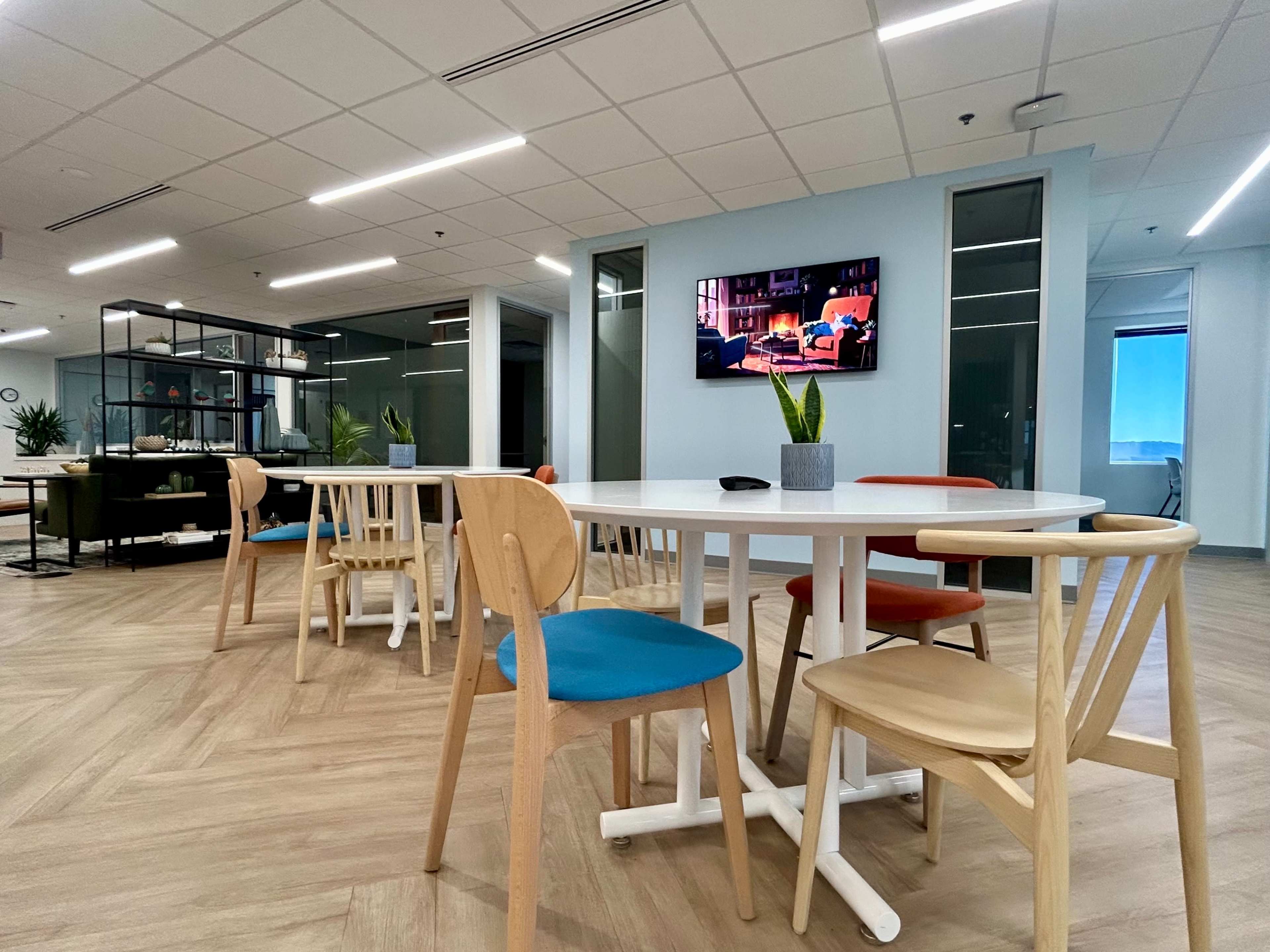 The image shows a modern office space featuring a circular table surrounded by colorful chairs, with a TV mounted on the wall displaying a cozy scene.