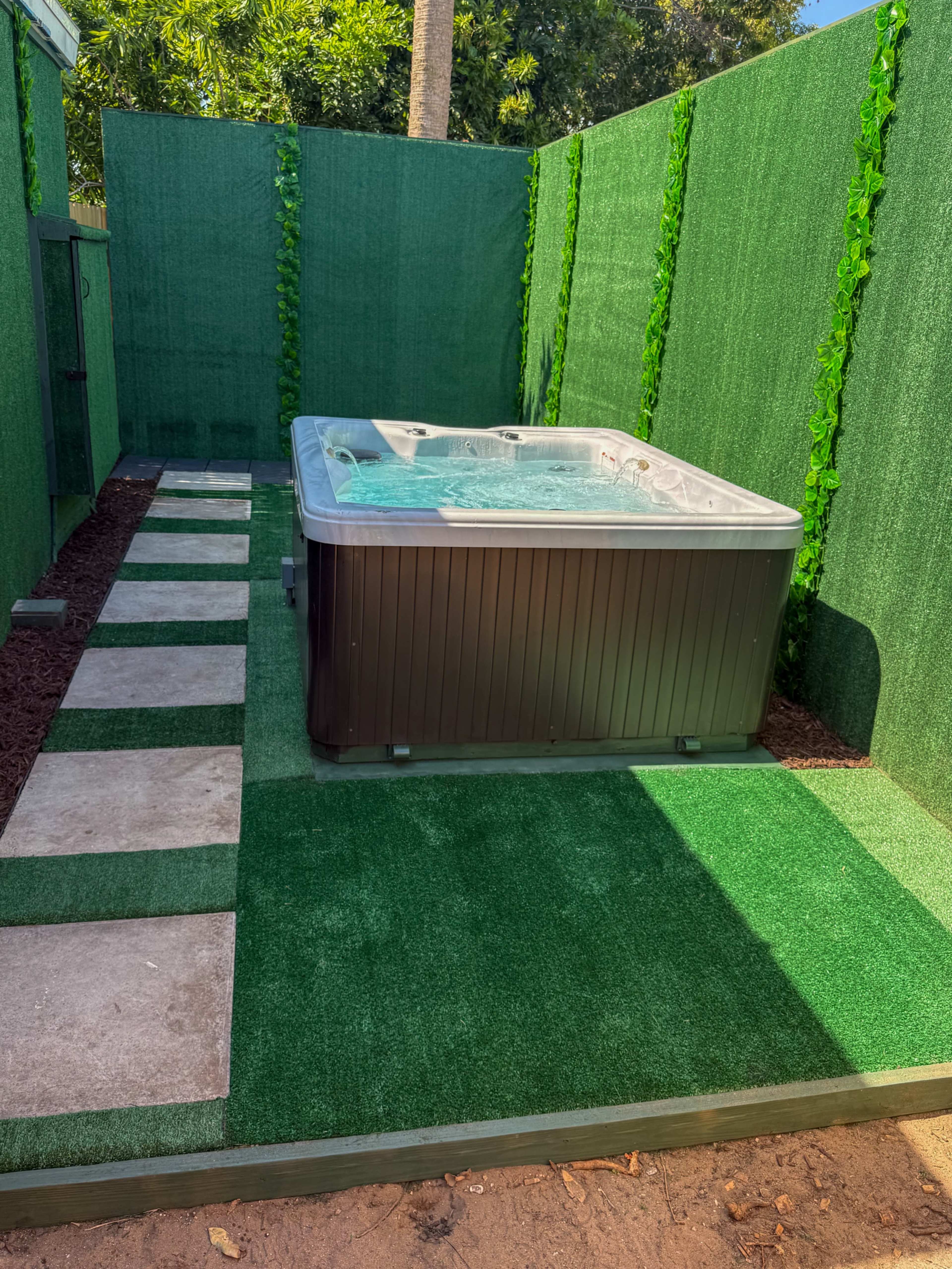 The image shows a hot tub surrounded by artificial grass and a green privacy wall, with a pathway made of stone slabs leading to it.