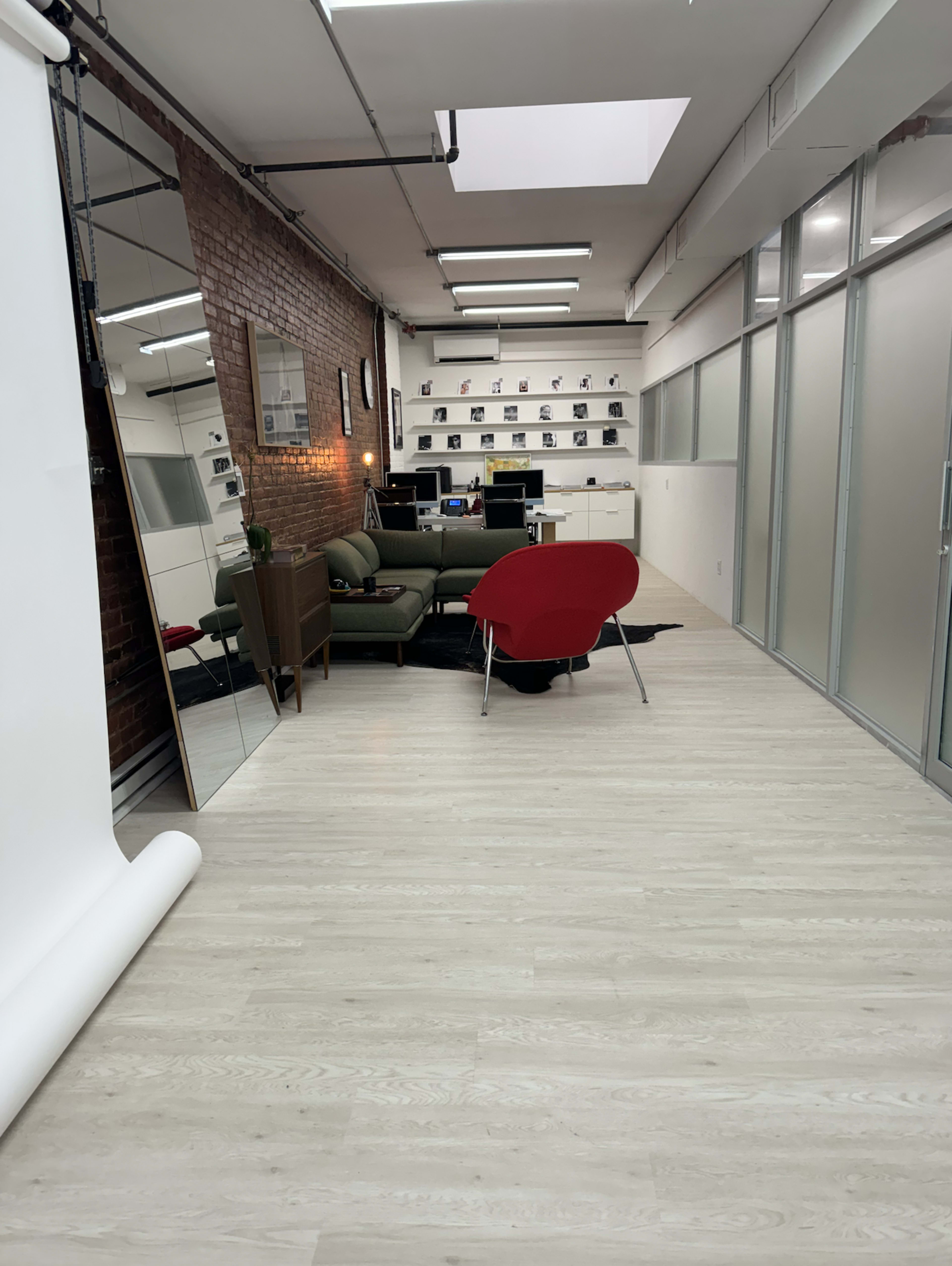 The image shows a well-lit modern workspace featuring a green sofa, a red chair, a large mirror, and multiple desks against a brick wall.