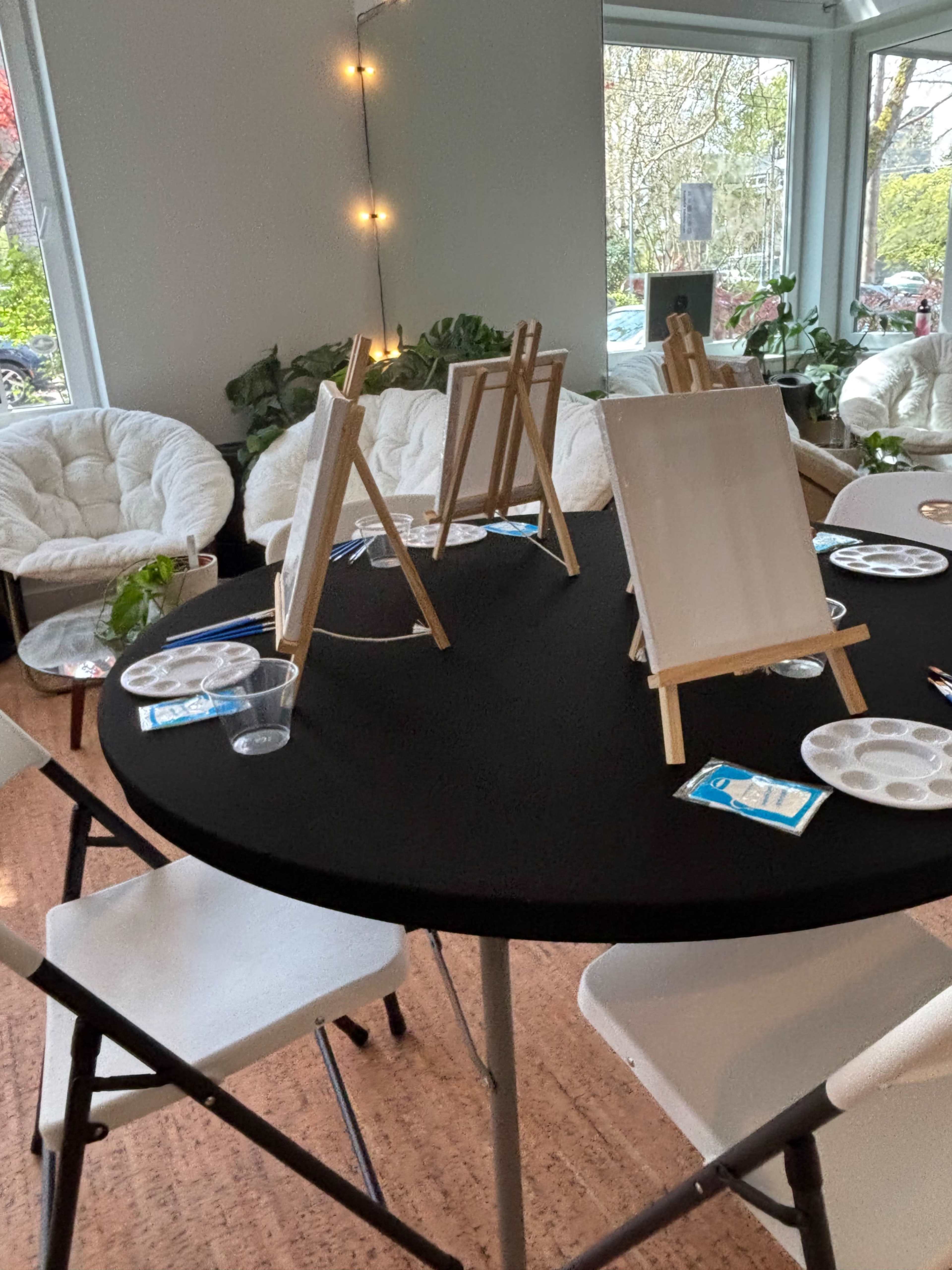 The image shows a circular table set up with blank canvases on easels, paint palettes, and plastic cups in a well-lit room with plants and comfortable chairs.