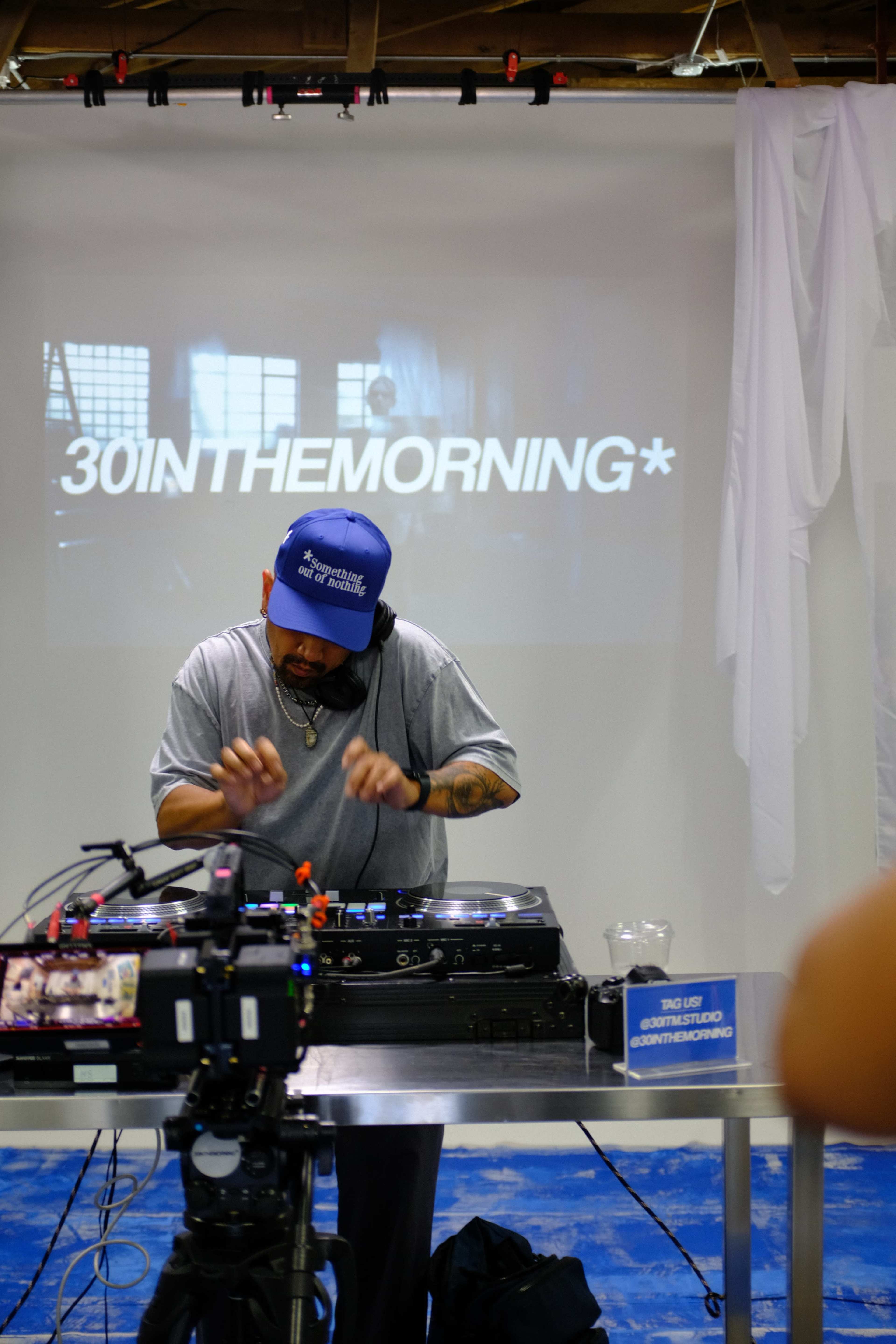 A man is performing as a DJ in front of a large screen displaying the text "30INTHEMORNING*" while equipment is set up on a table.