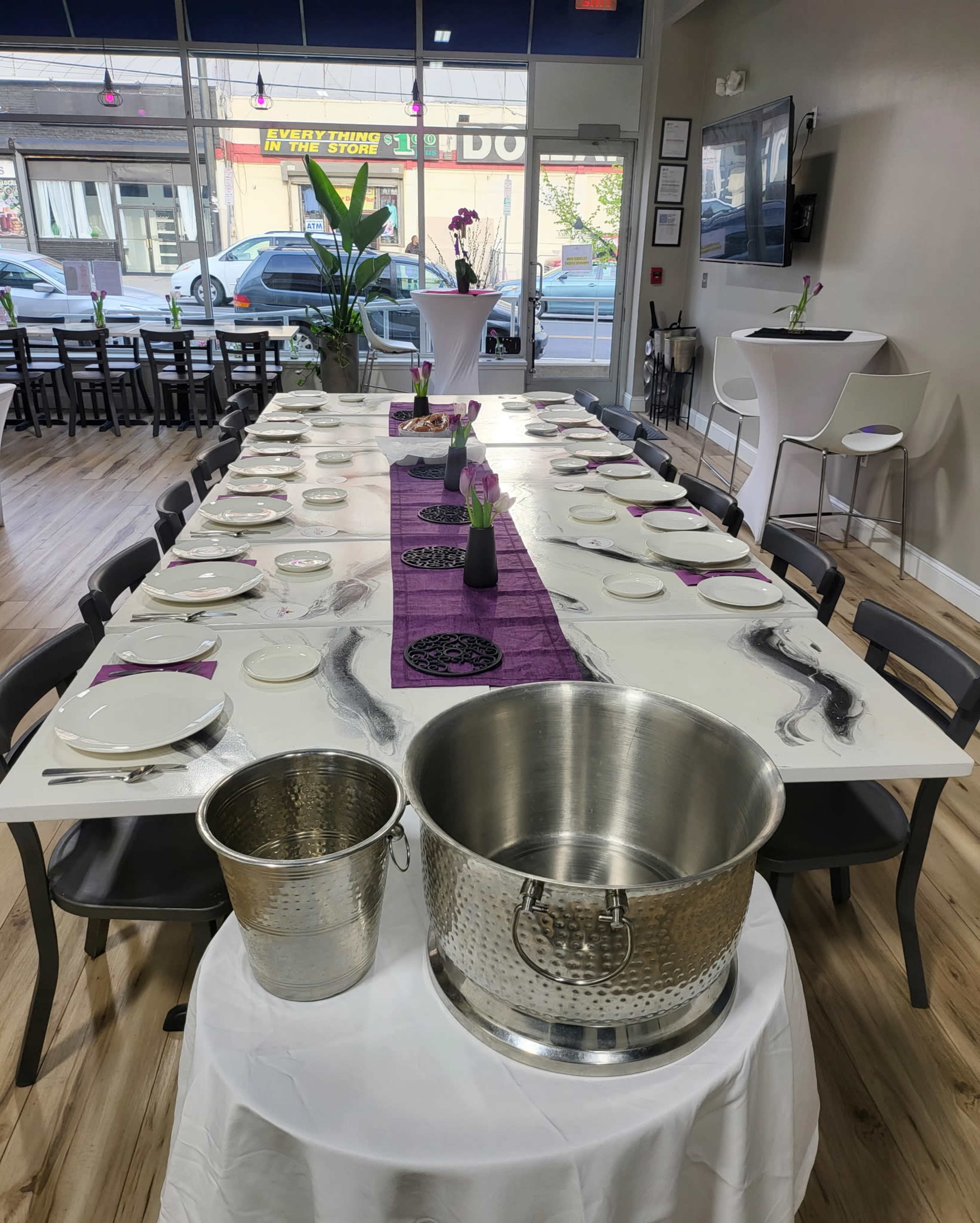 A long dining table is set with plates and cutlery, featuring a purple table runner and two silver ice buckets at one end, with a restaurant interior visible in the background.