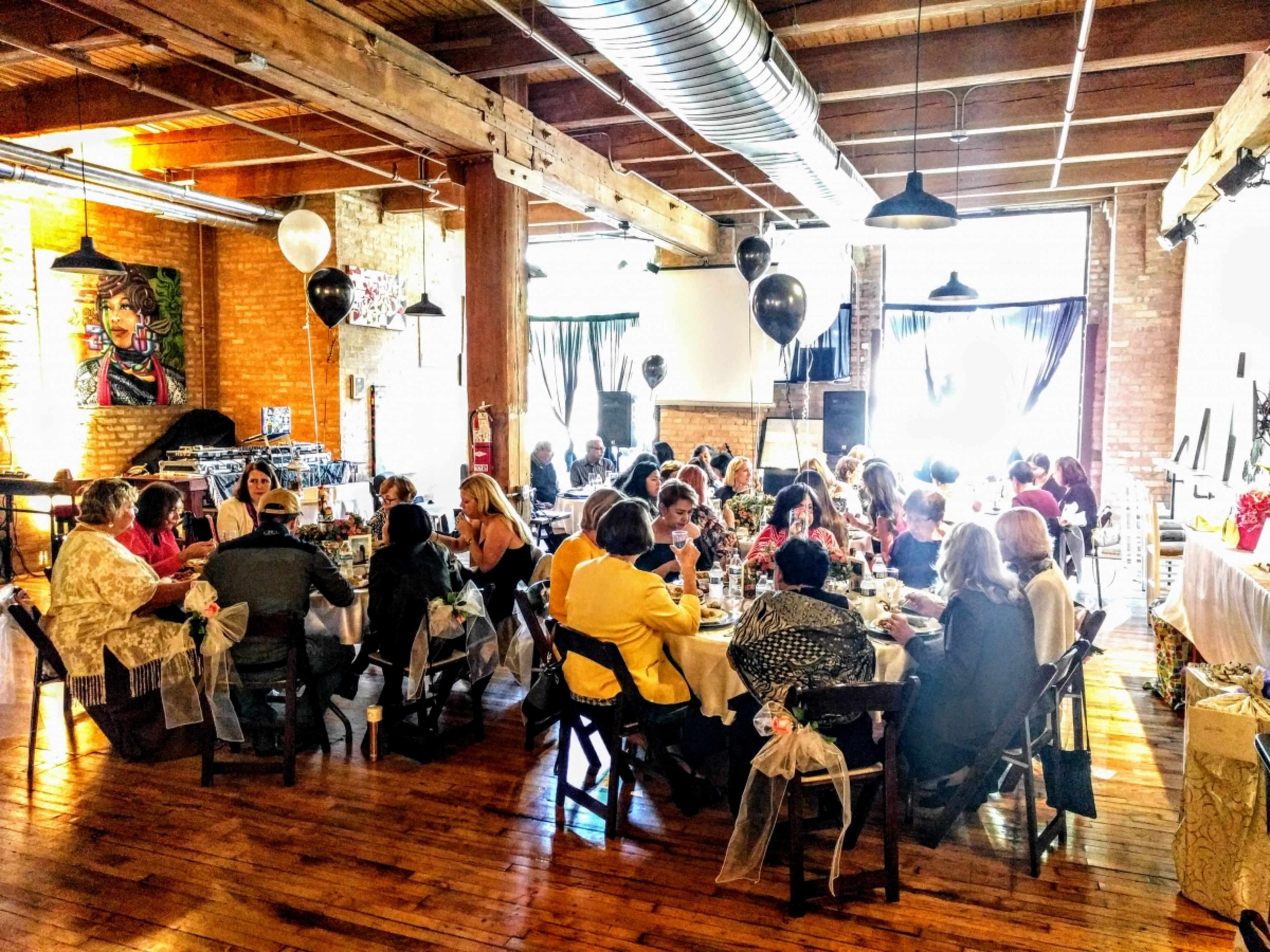 A group of women gathers around tables in a spacious, well-lit room decorated with artwork and balloons.