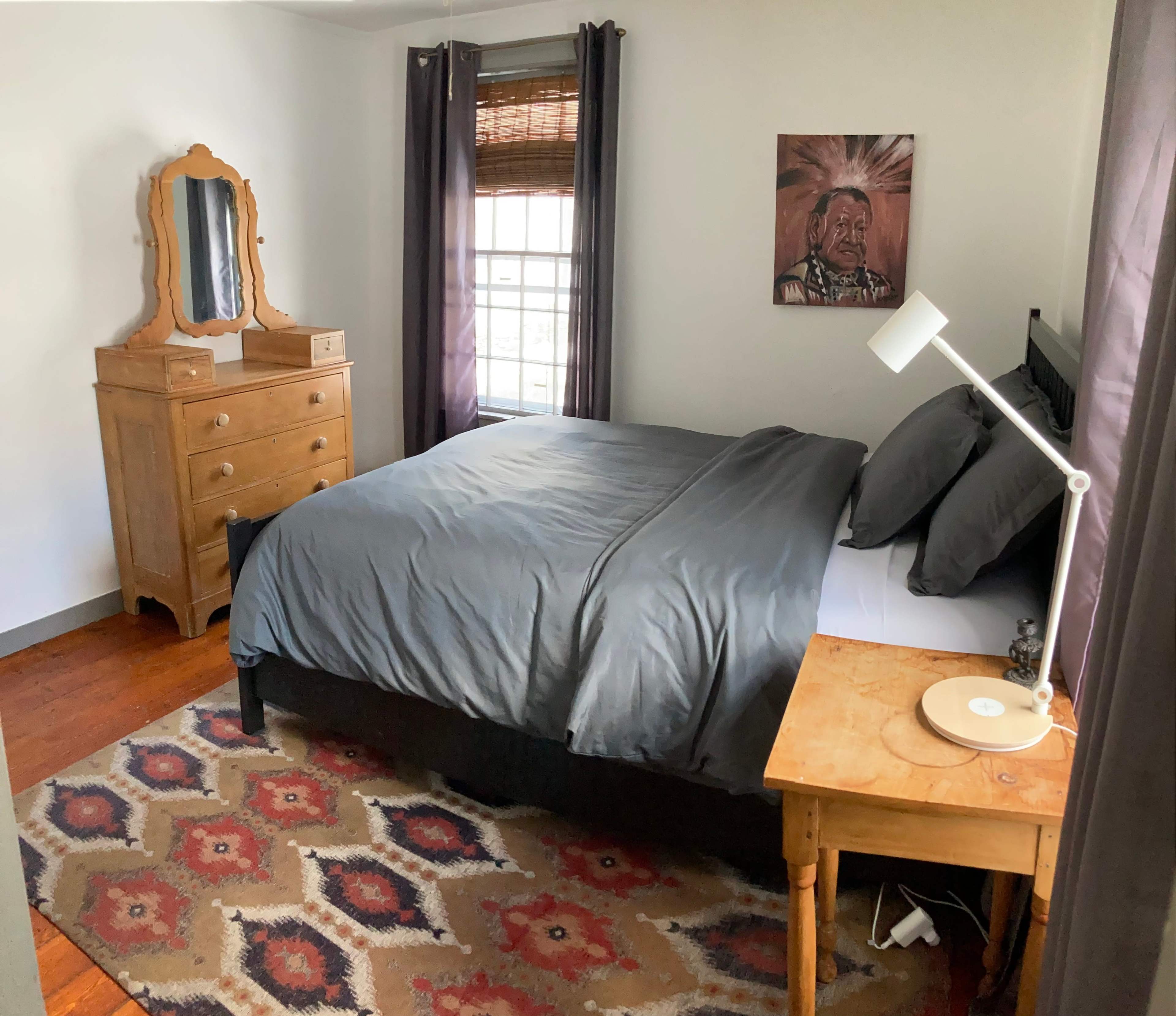 The scene shows a bedroom featuring a bed with gray bedding, a wooden dresser and mirror, a side table with a lamp, and a patterned rug on the floor.
