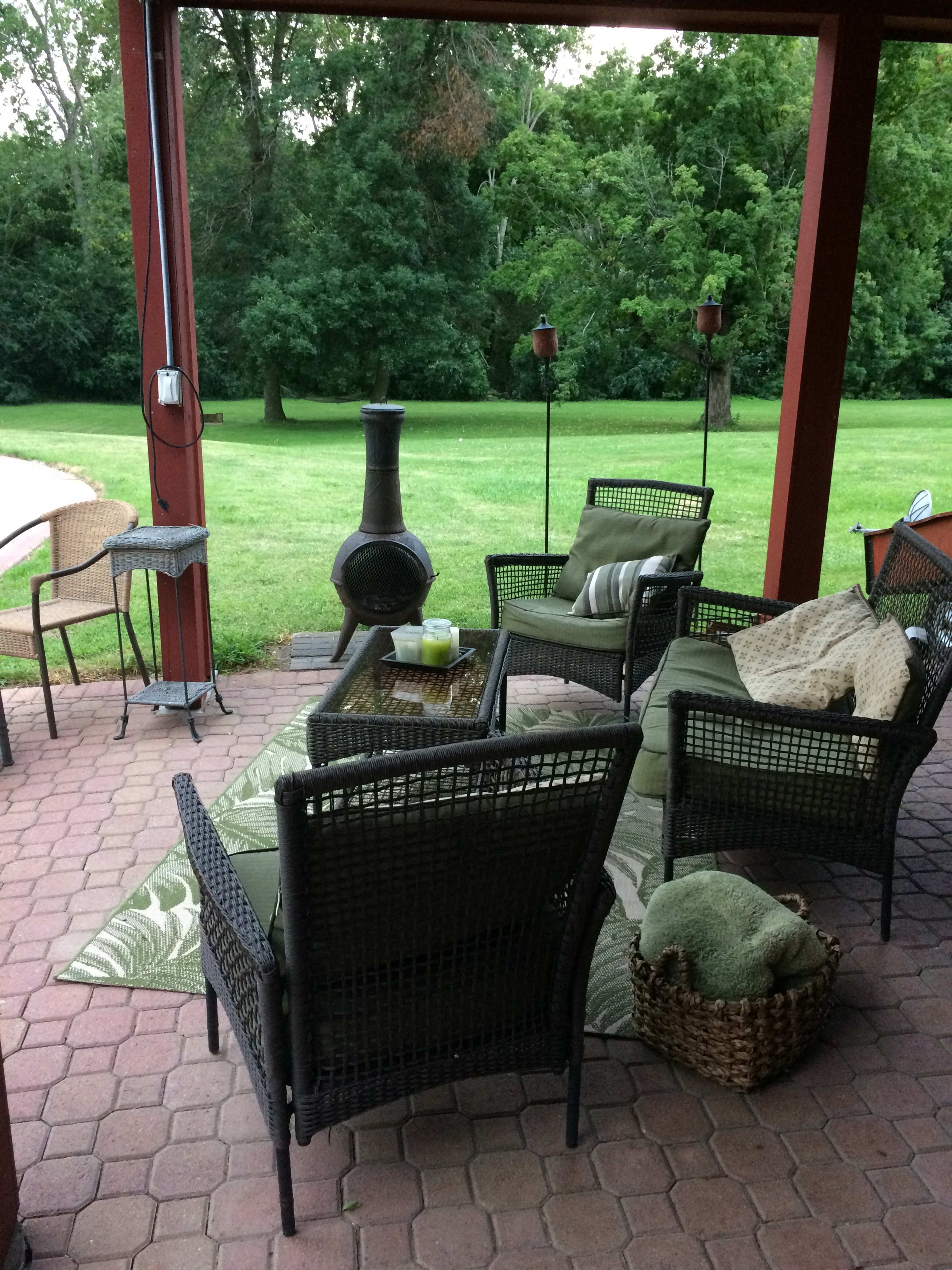 A cozy outdoor seating area features wicker chairs with cushions, a table, and a chiminea, set against a backdrop of lush greenery.