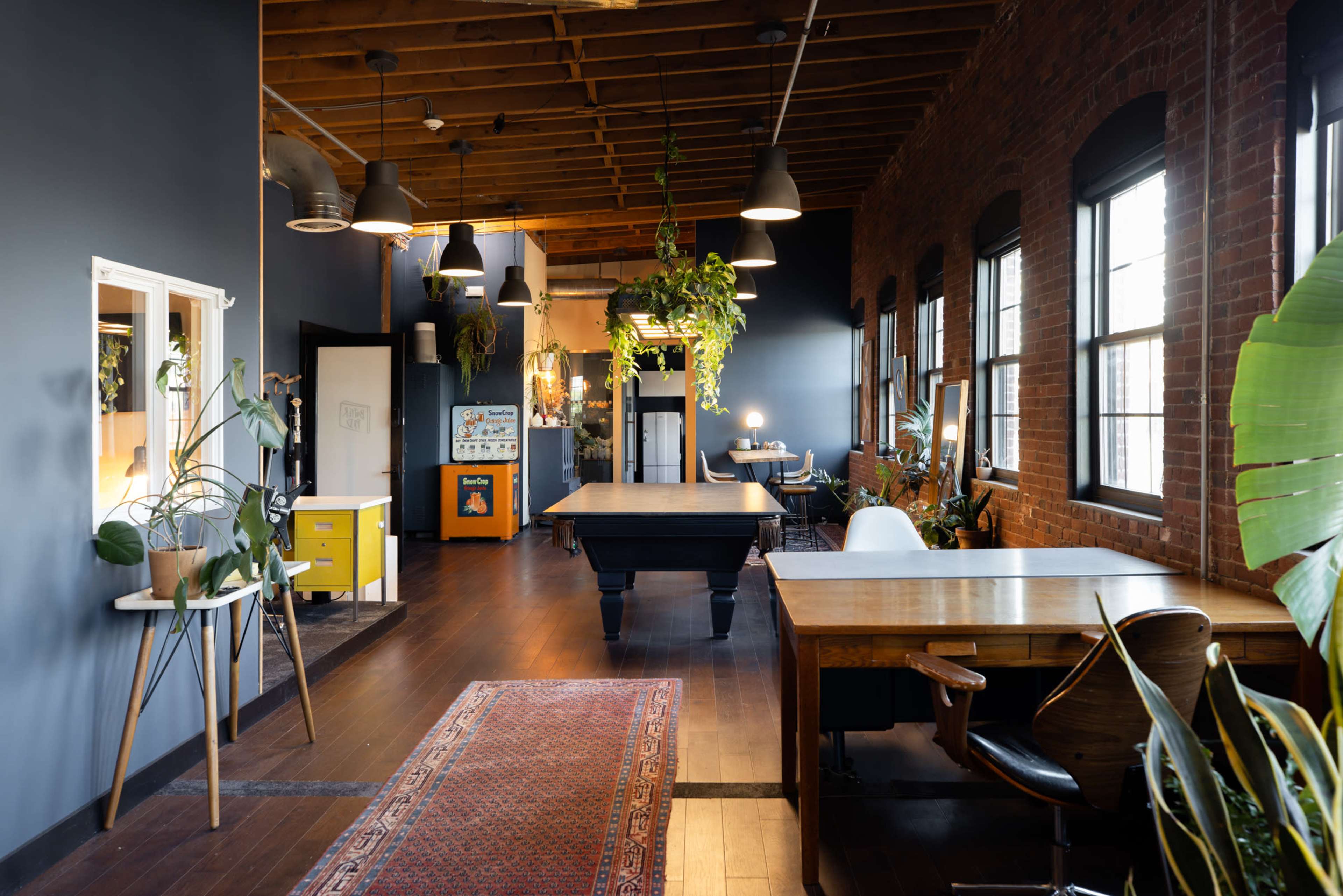 The image shows a spacious office area with wooden flooring, large windows, and various desks arranged along the walls, adorned with plants and decorative elements.