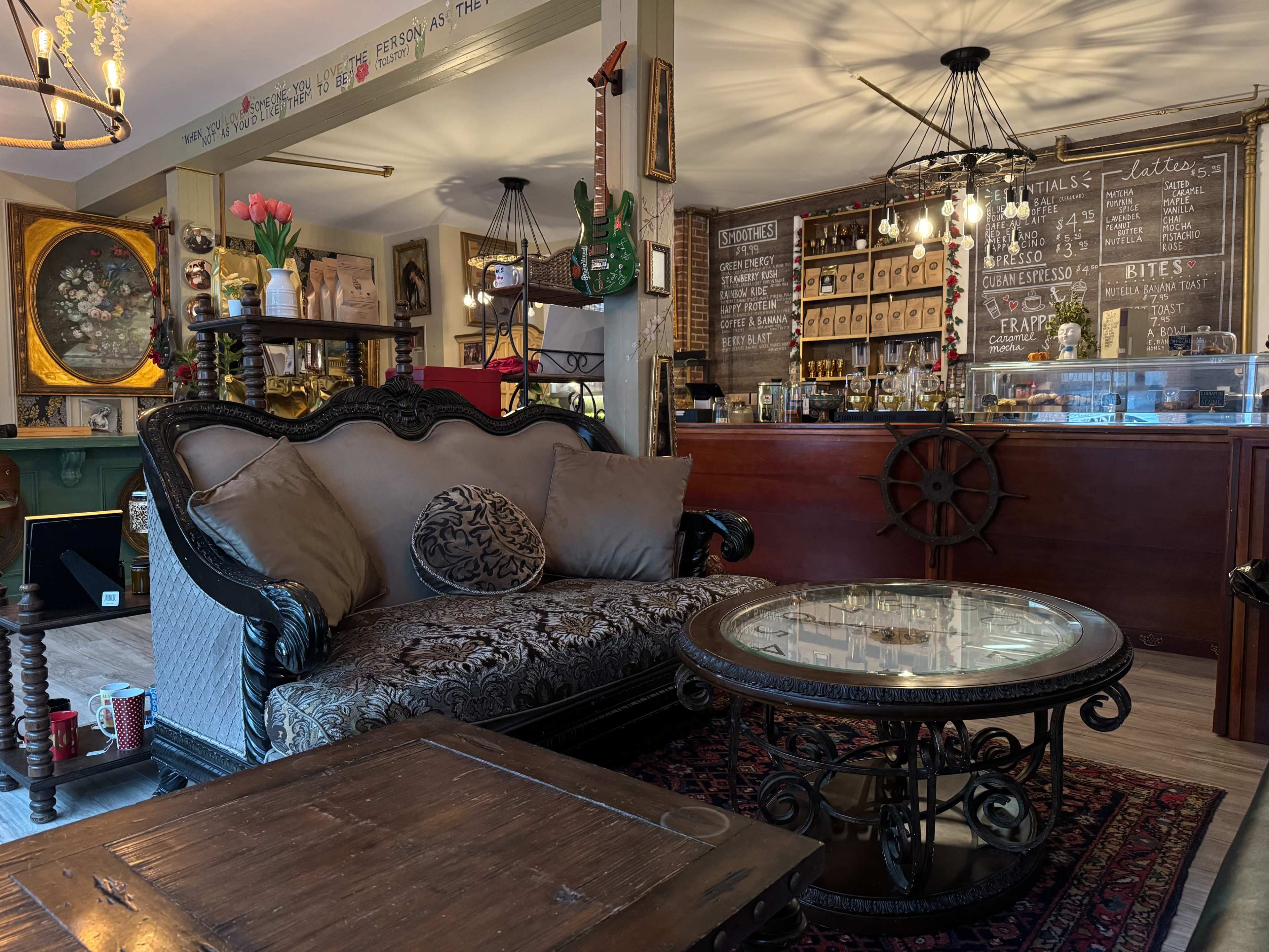 The image shows a cozy café interior featuring a vintage-style couch, a round glass coffee table, and a counter with a menu board in the background.
