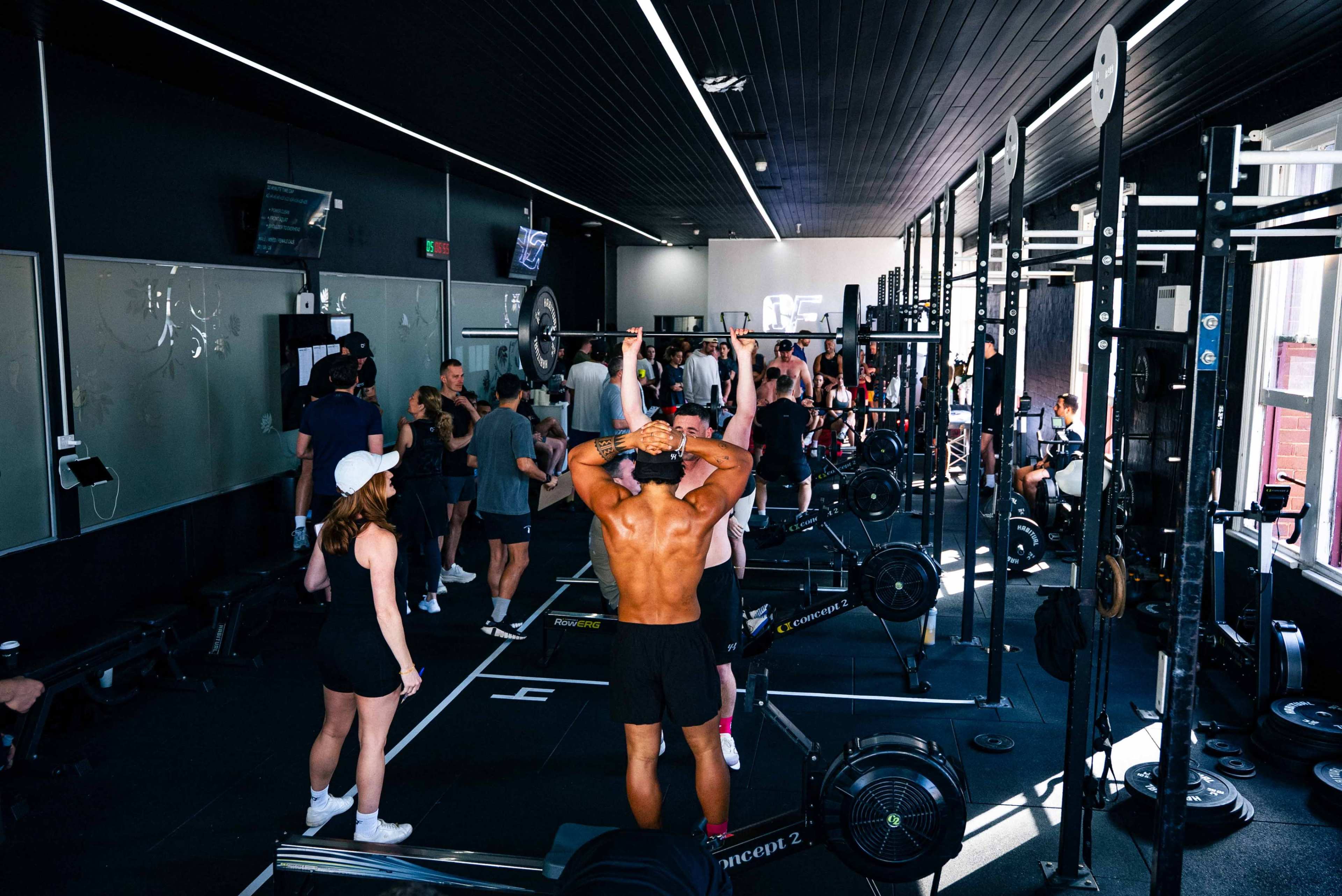 A crowded gym features various individuals working out with weights and exercise equipment.