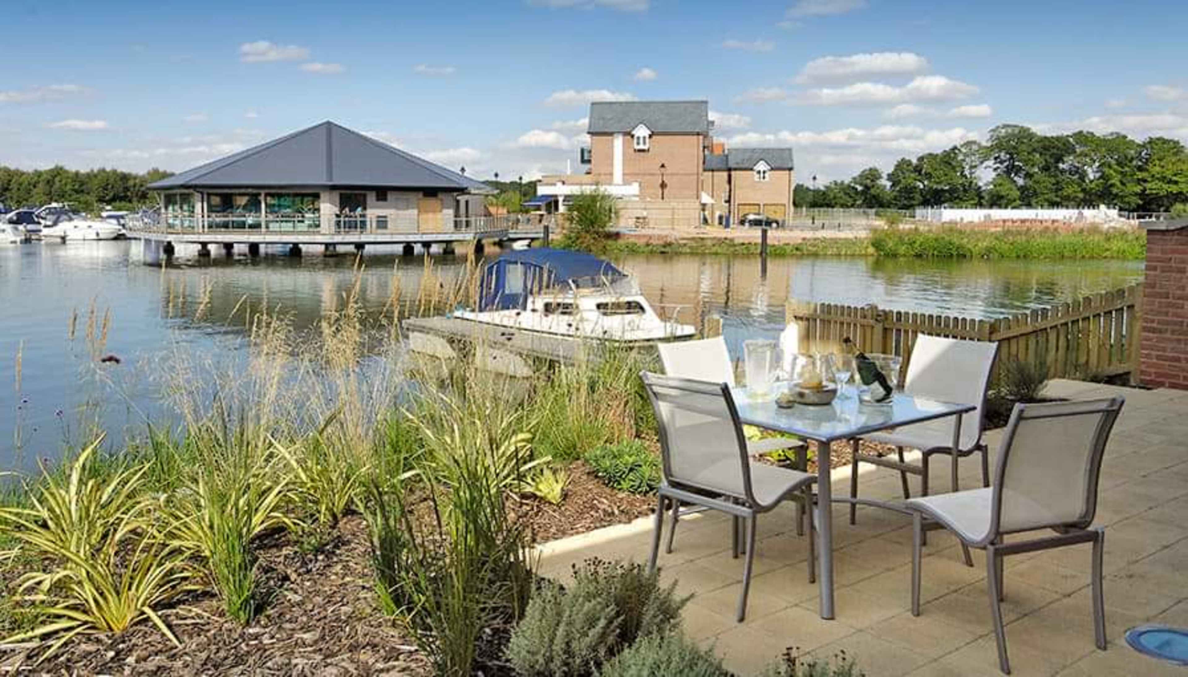 The scene shows a waterfront dining area with a table set for a meal, overlooking a marina with boats and a building in the background.