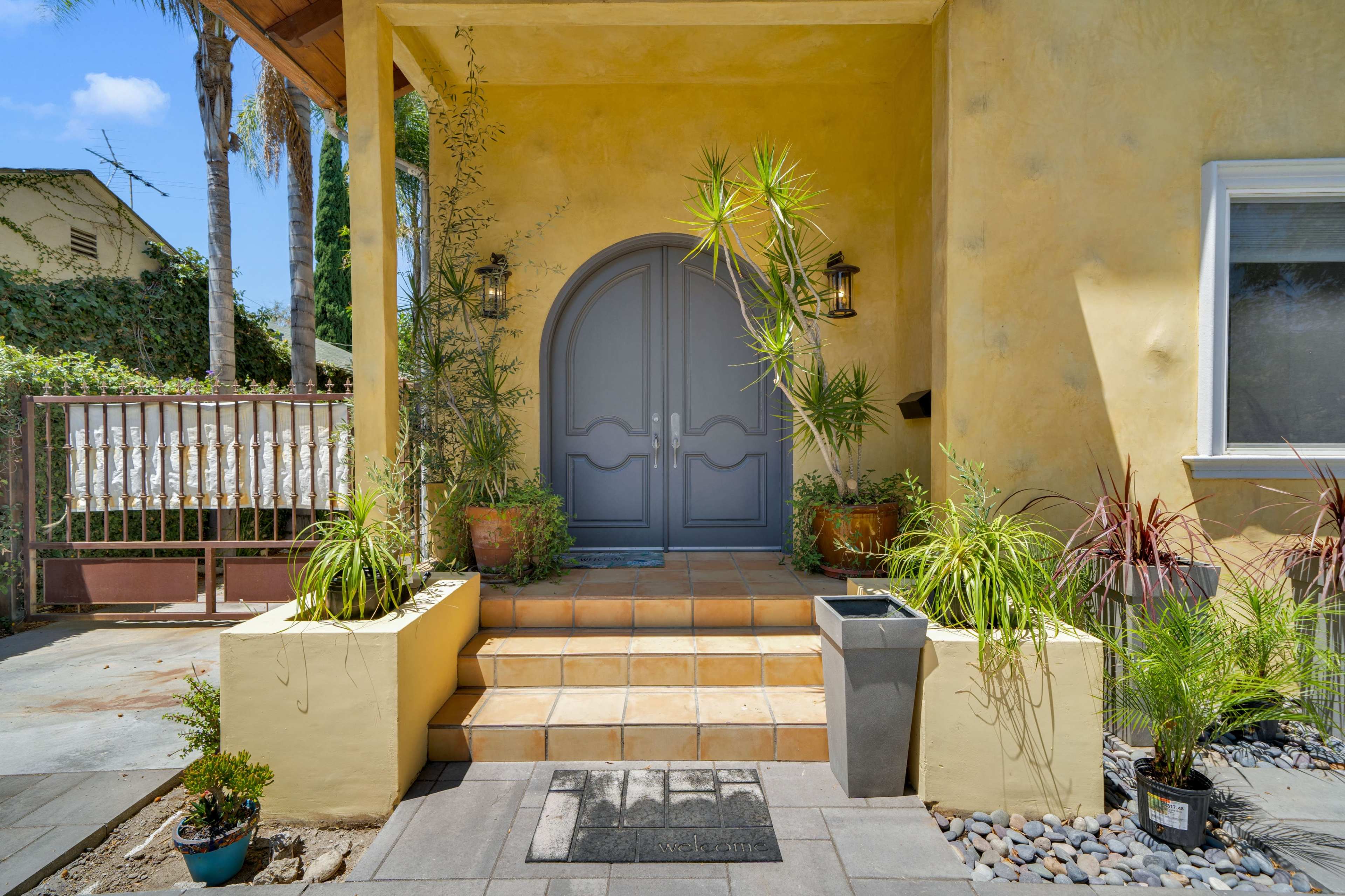 The image shows a yellow-walled entrance with a set of double doors, flanked by potted plants, leading up to a tiled staircase.