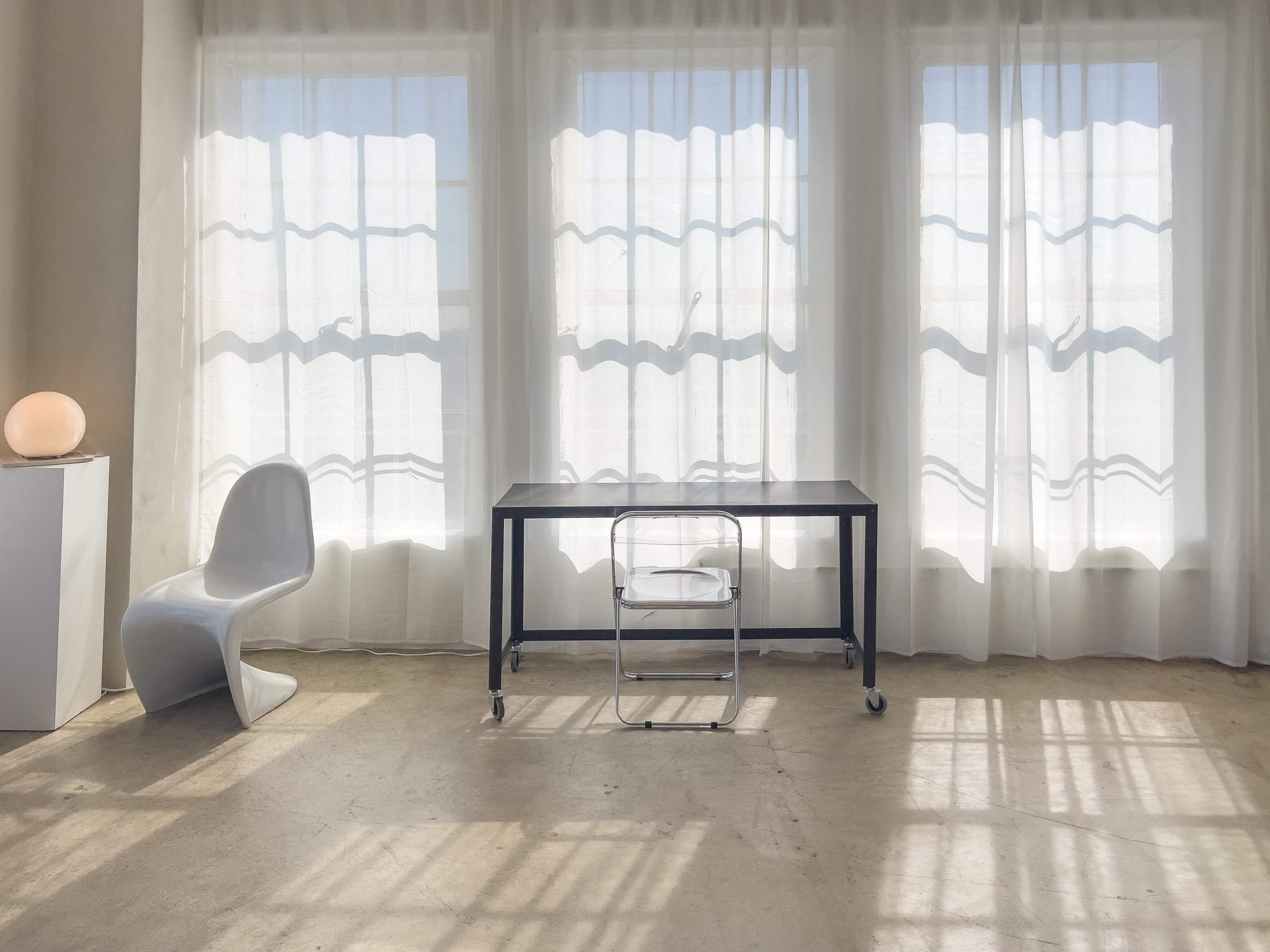 A minimalistic room features a table with a wheeled chair positioned between large, bright windows framed by sheer white curtains.