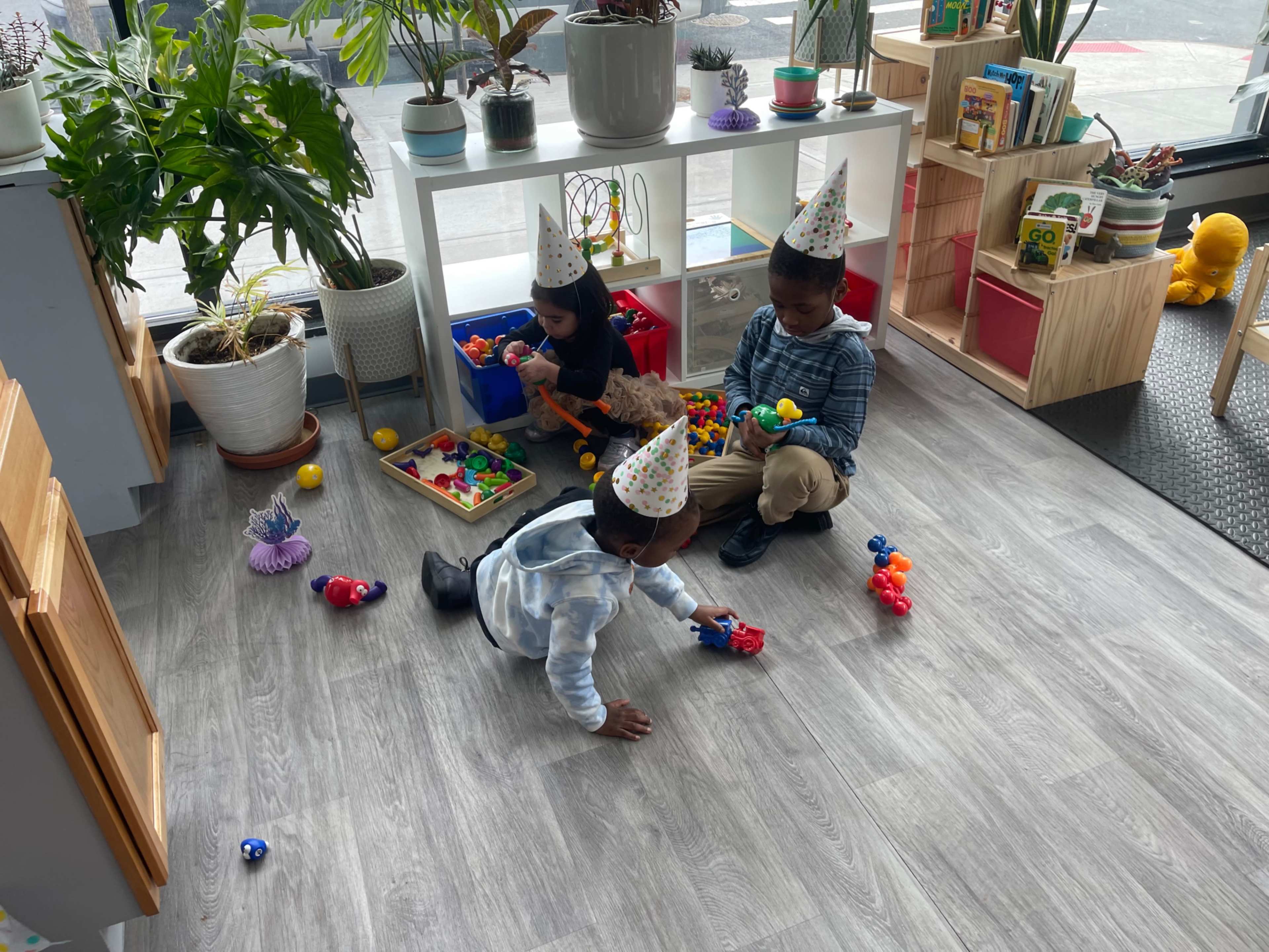 Three children play on a wooden floor in a bright room filled with plants and educational toys, while wearing party hats.