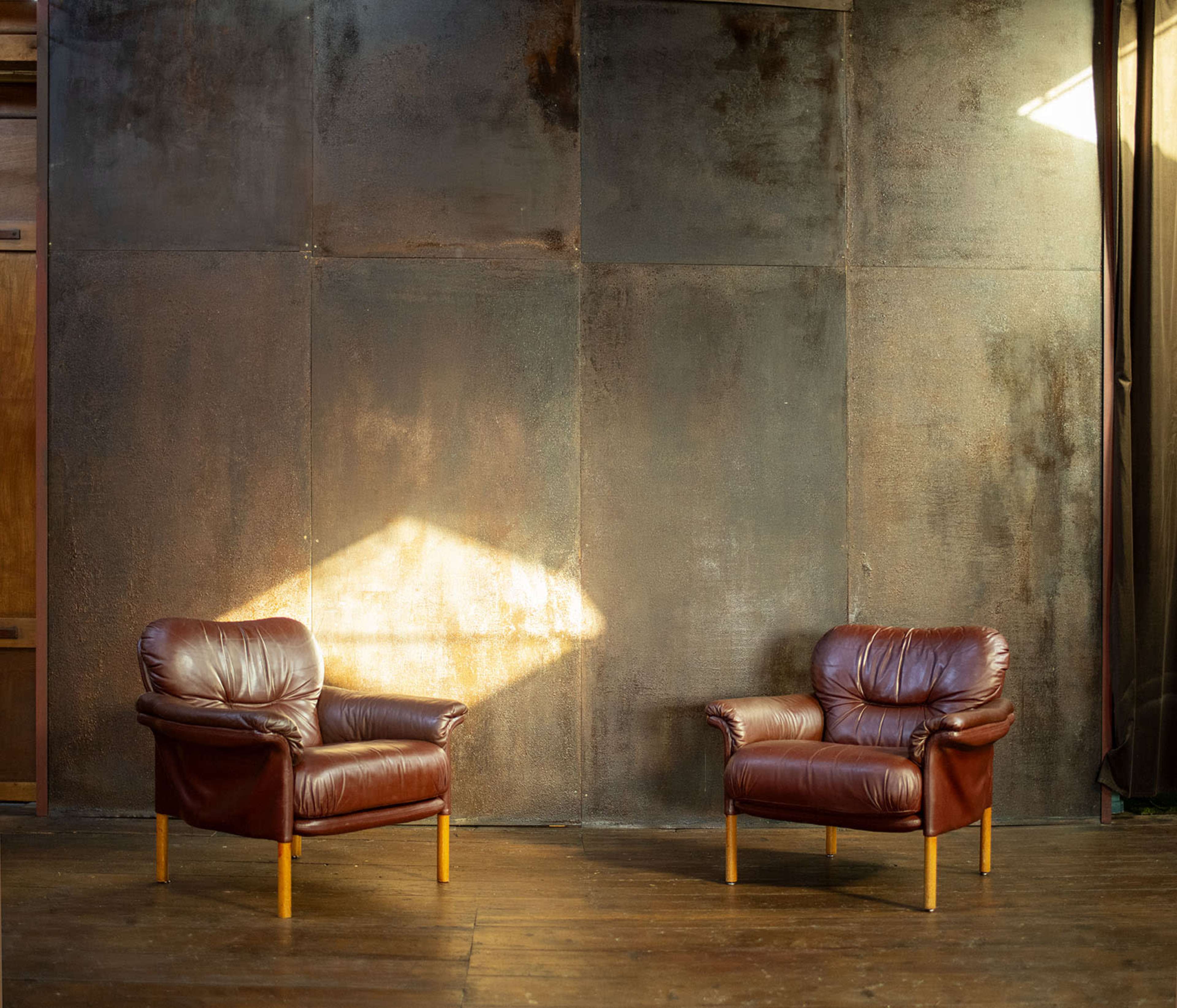 Two brown leather chairs are positioned facing each other against a textured metal wall, with sunlight illuminating one chair.