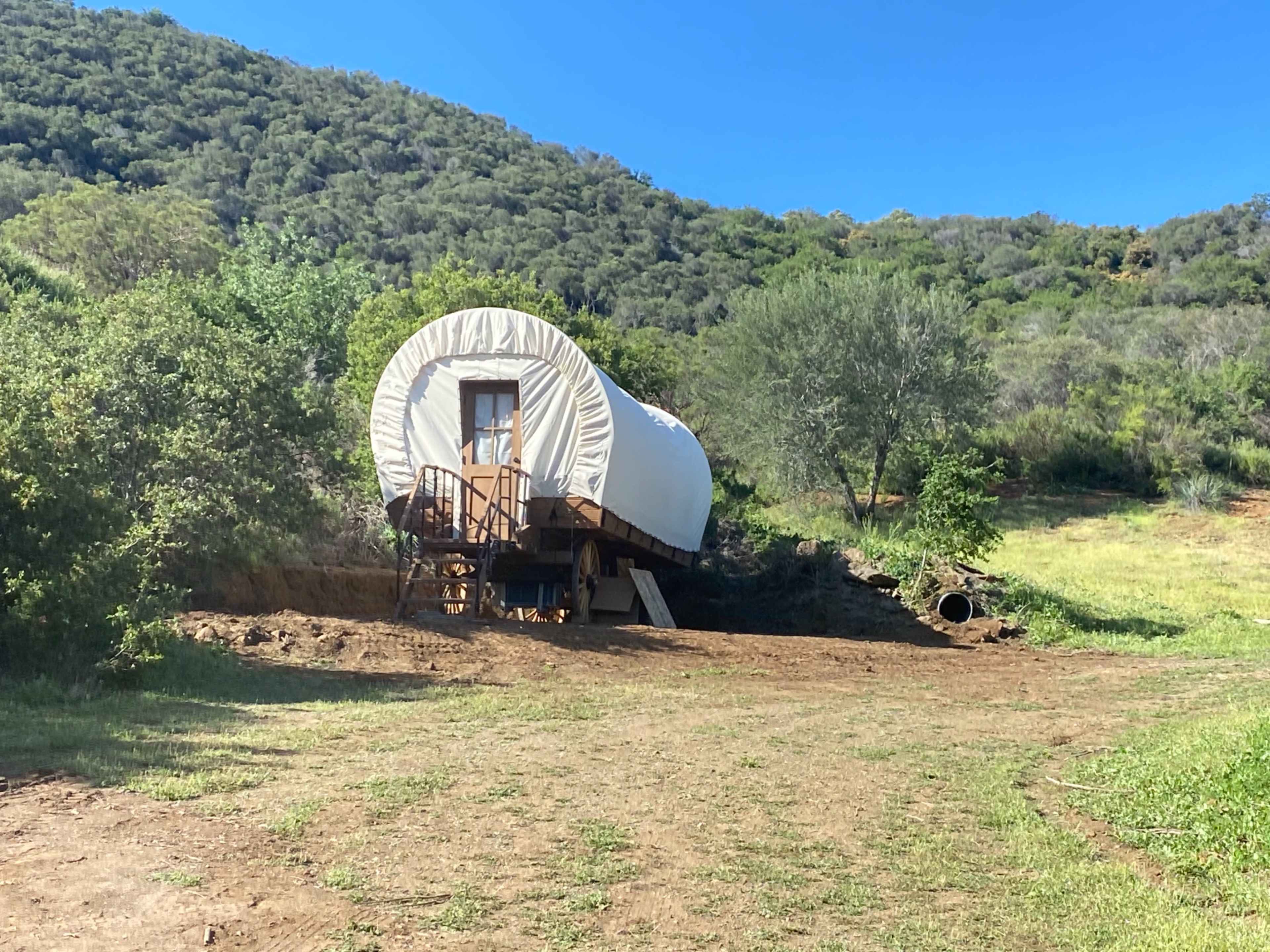 A covered wagon is positioned on a small hill in a grassy area, surrounded by trees and hills.