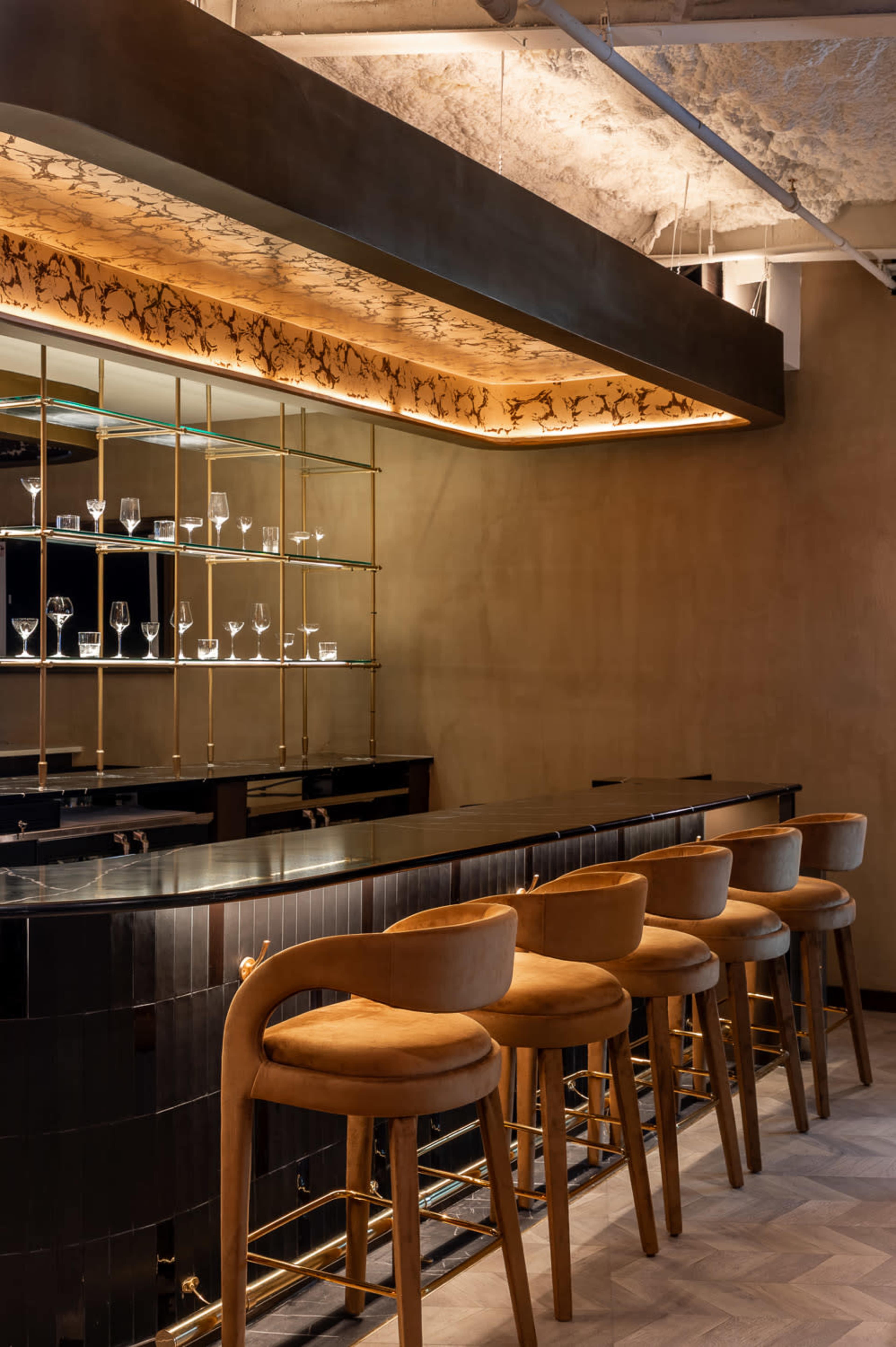 The image shows a modern bar area with a sleek black counter, elegant high stools, and illuminated shelves displaying glassware.
