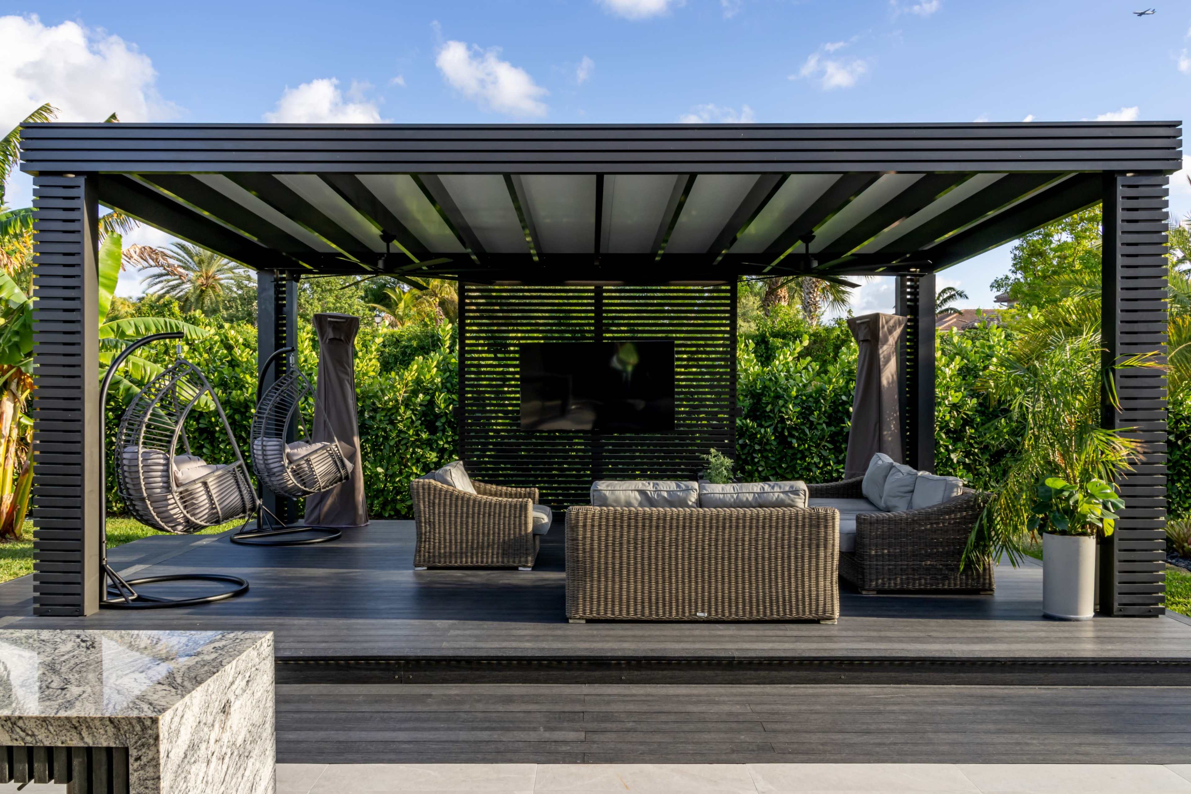 A modern outdoor patio features a black pergola with comfortable seating, hanging chairs, and a mounted television, surrounded by lush greenery.