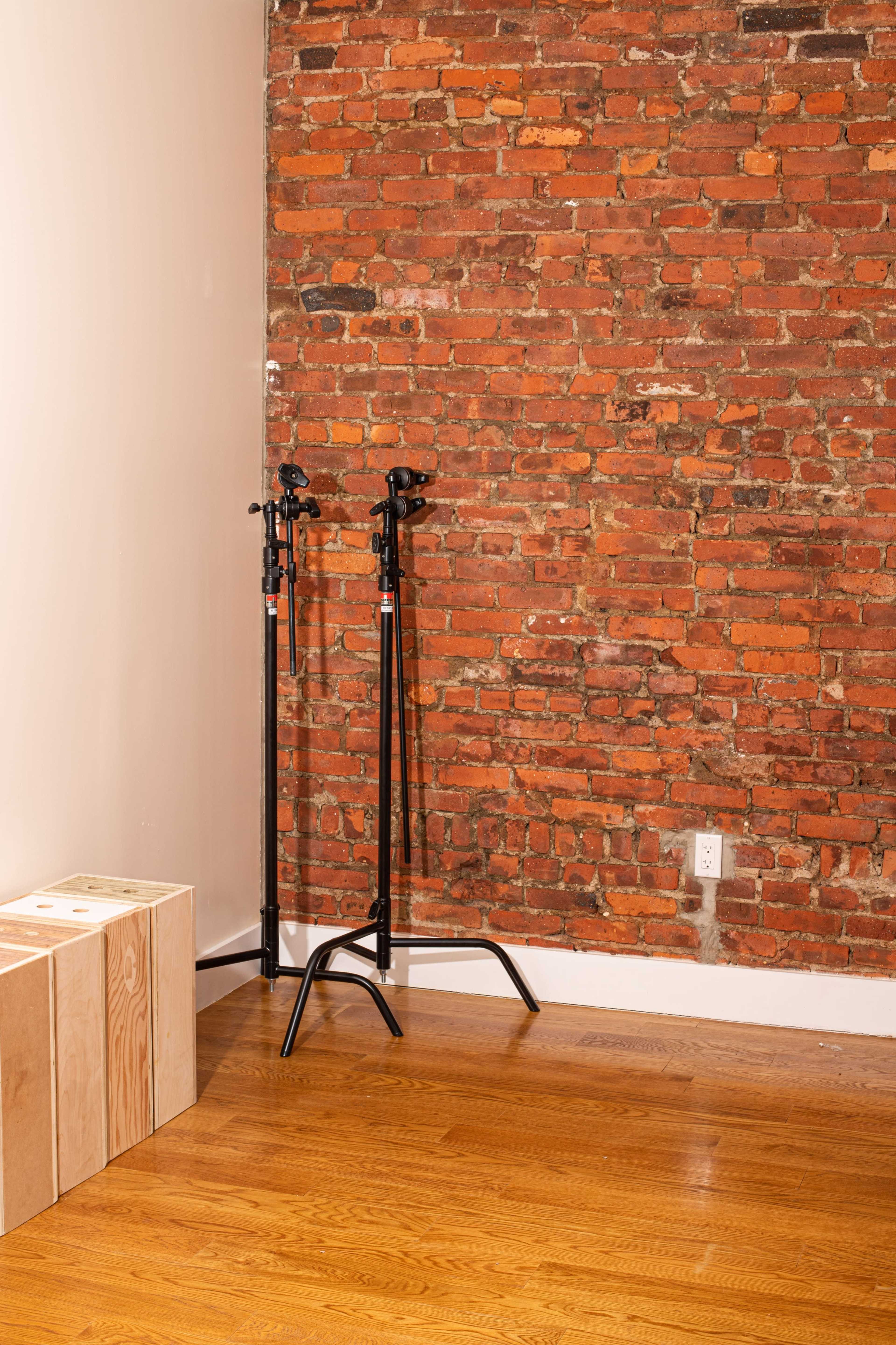 Two black stands positioned next to a rough, red-brick wall in a room with wooden flooring.