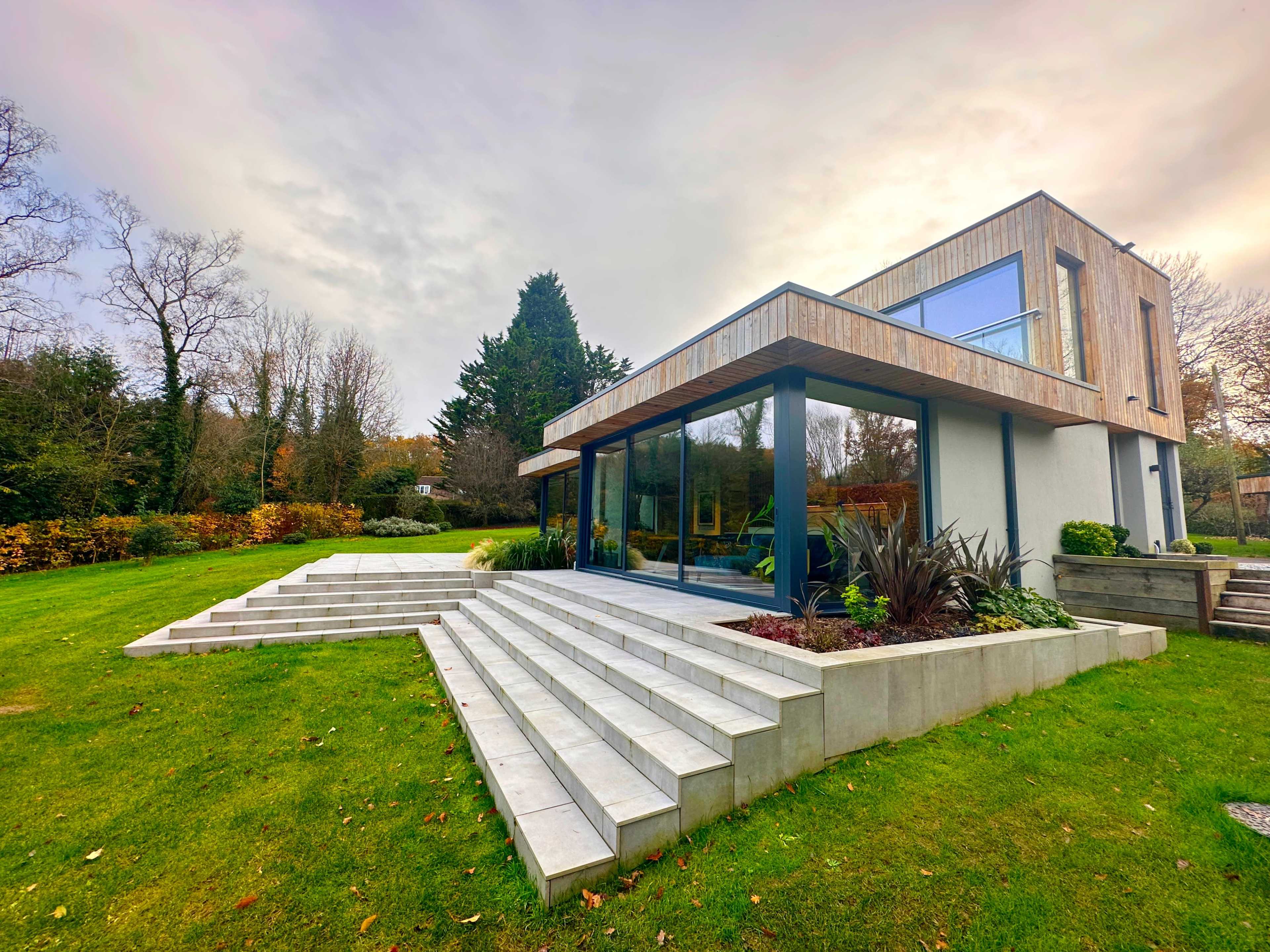 A modern house with large glass windows and a wooden overhang, surrounded by a well-manicured lawn and landscaped gardens.