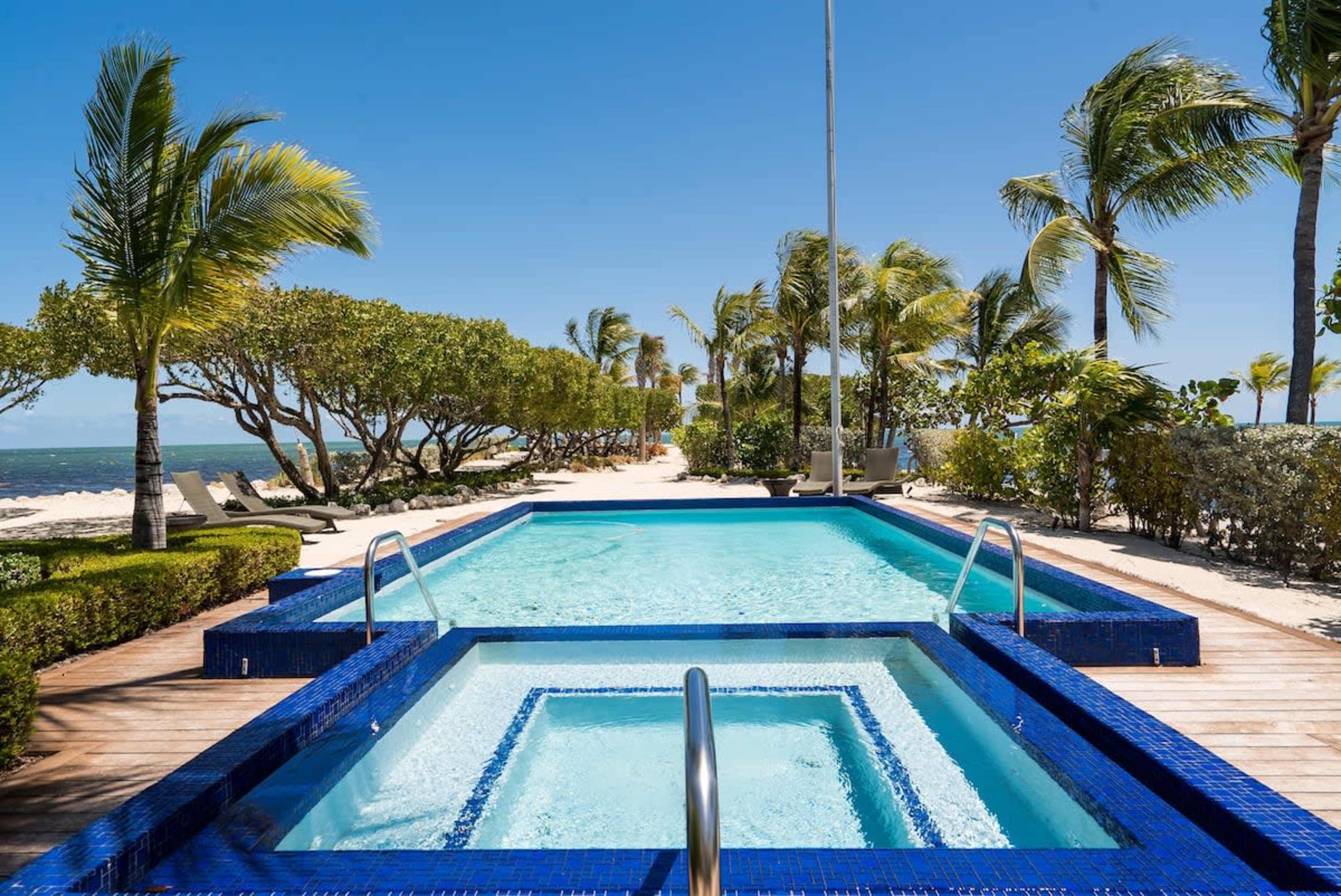 The image shows a blue-tiled swimming pool and hot tub surrounded by palm trees and landscaped greenery, overlooking a beach.