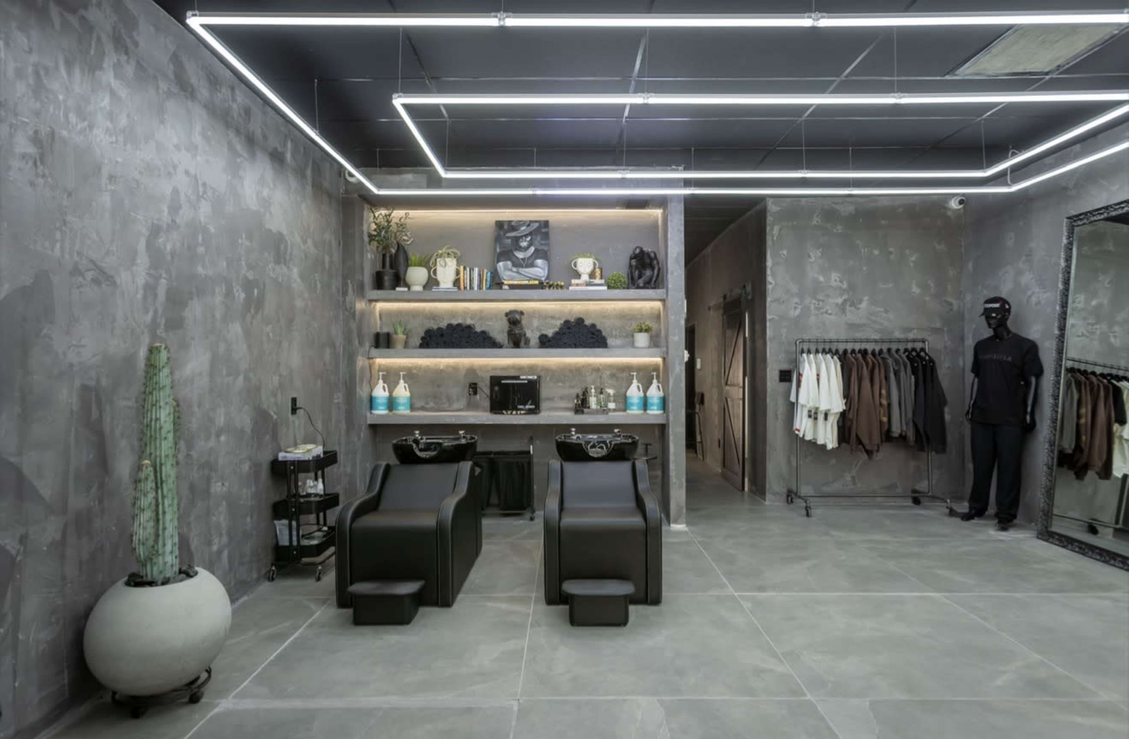 The image shows a modern salon interior with two black shampoo stations, a clothing rack, and minimalist decor featuring gray walls and a cactus.