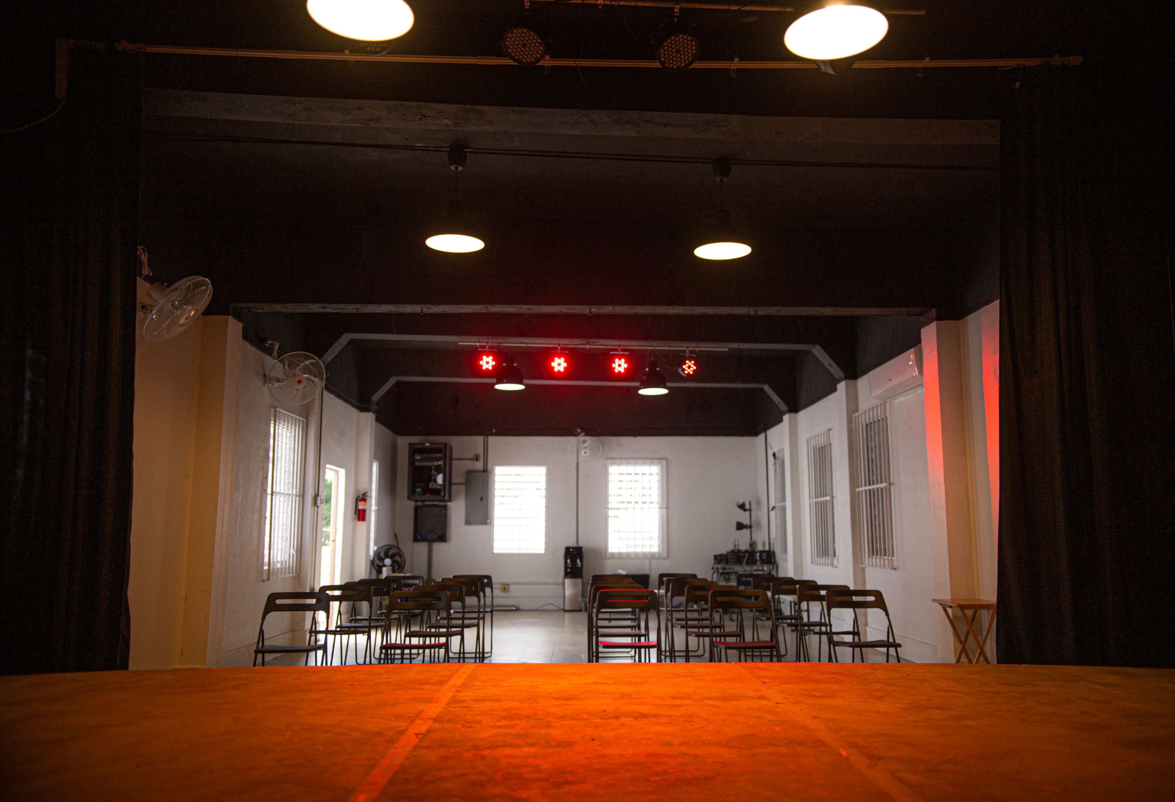 The image shows an empty auditorium with rows of chairs facing a stage, illuminated by overhead lights.