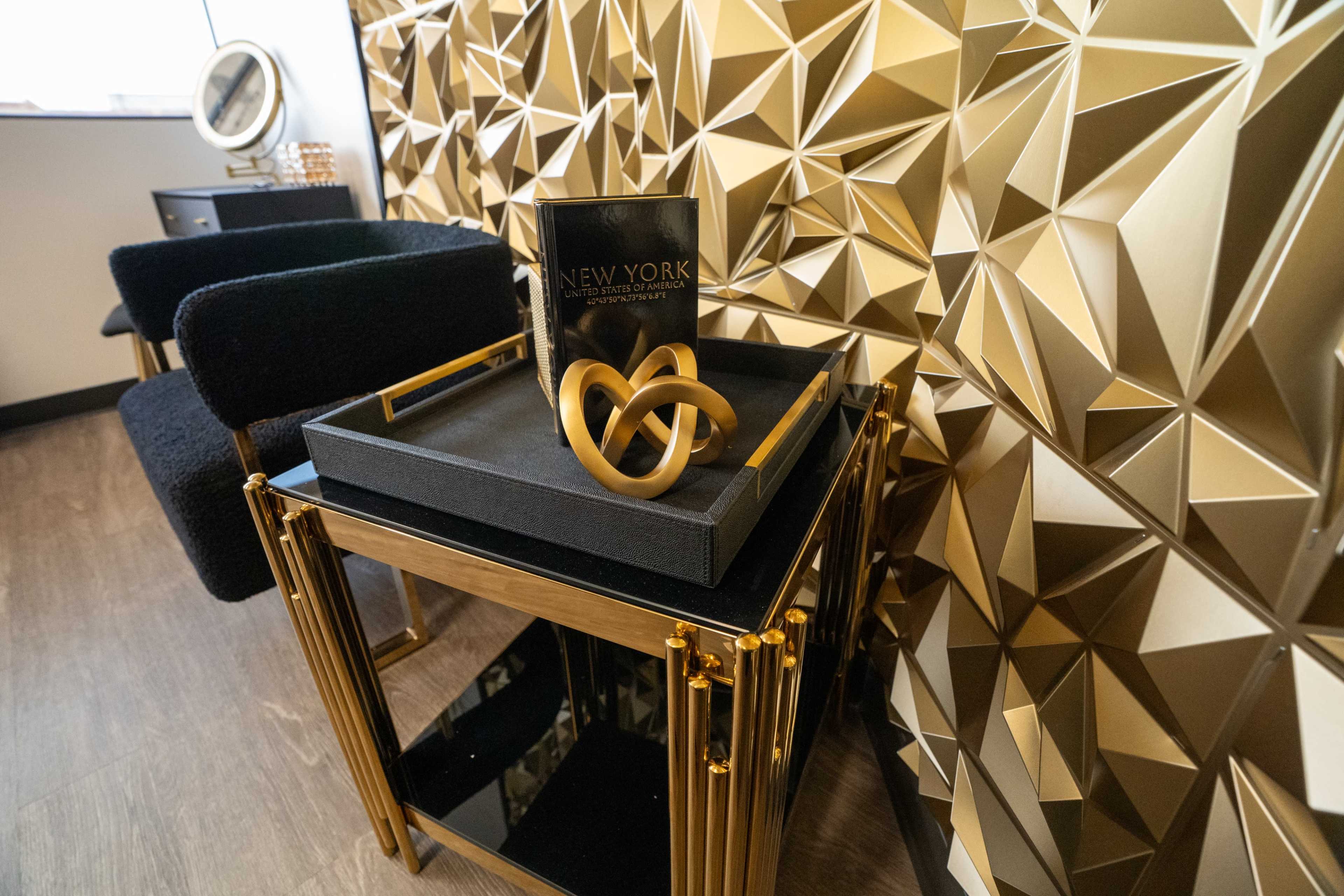 A small black and gold side table displays a decorative tray with a twisted gold sculpture and a book titled "NEW YORK" in a stylish room featuring a geometric gold wall.
