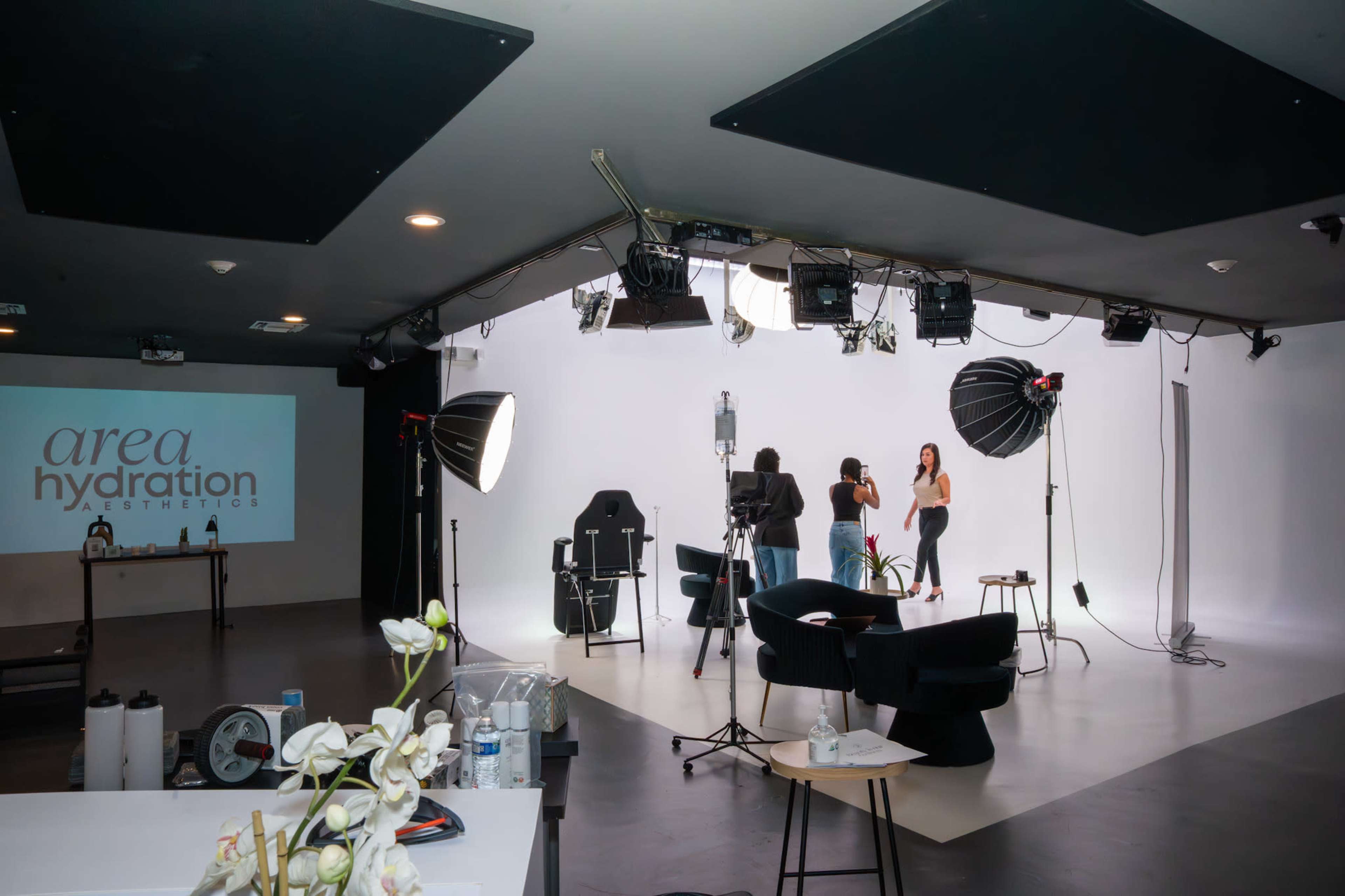 A photography studio with a white backdrop features two models being filmed, while lighting equipment and props are arranged around the space.