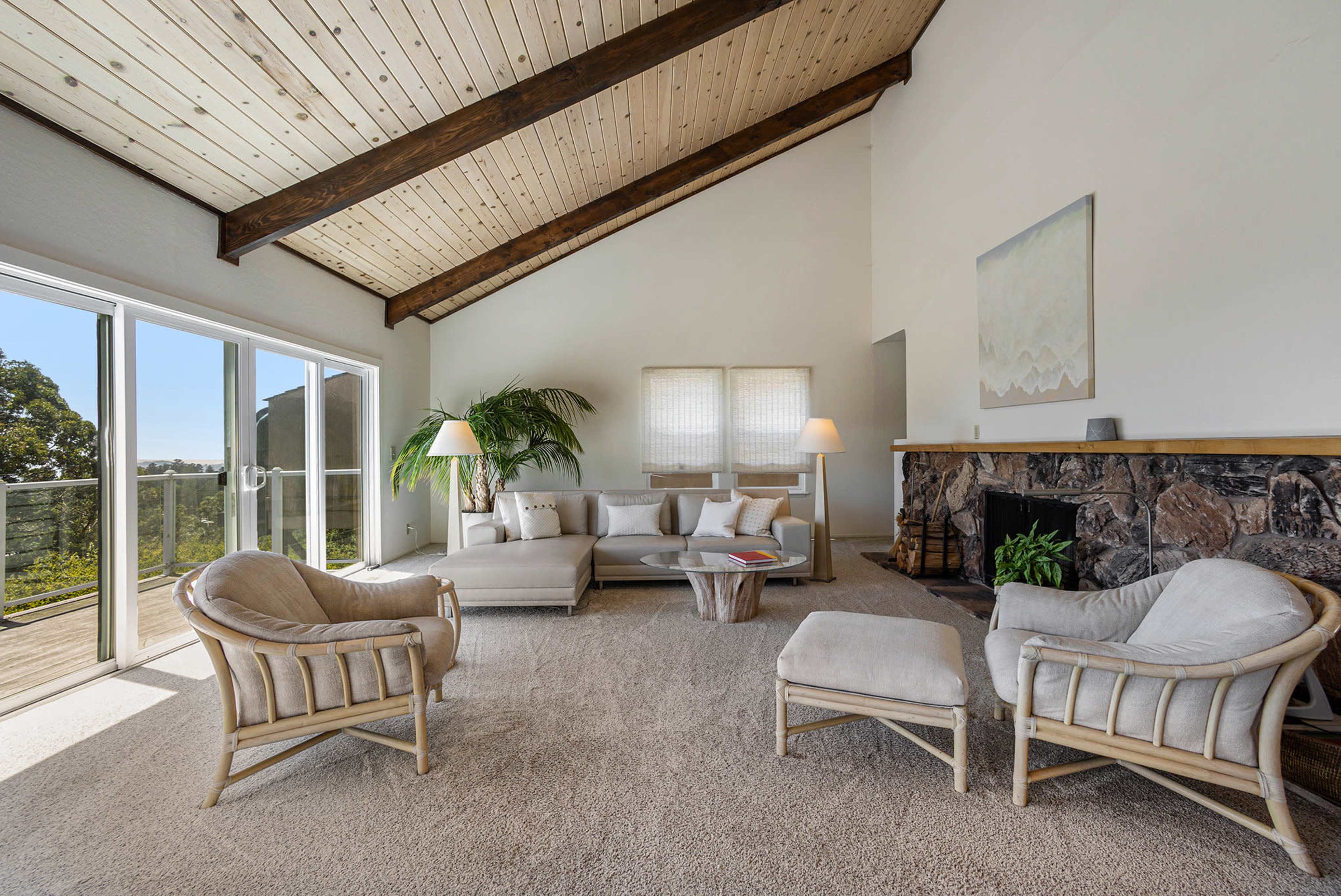 A spacious living room features high wooden ceilings, large windows with sliding doors, and a simple seating area with two armchairs and a sofa arranged around a coffee table.