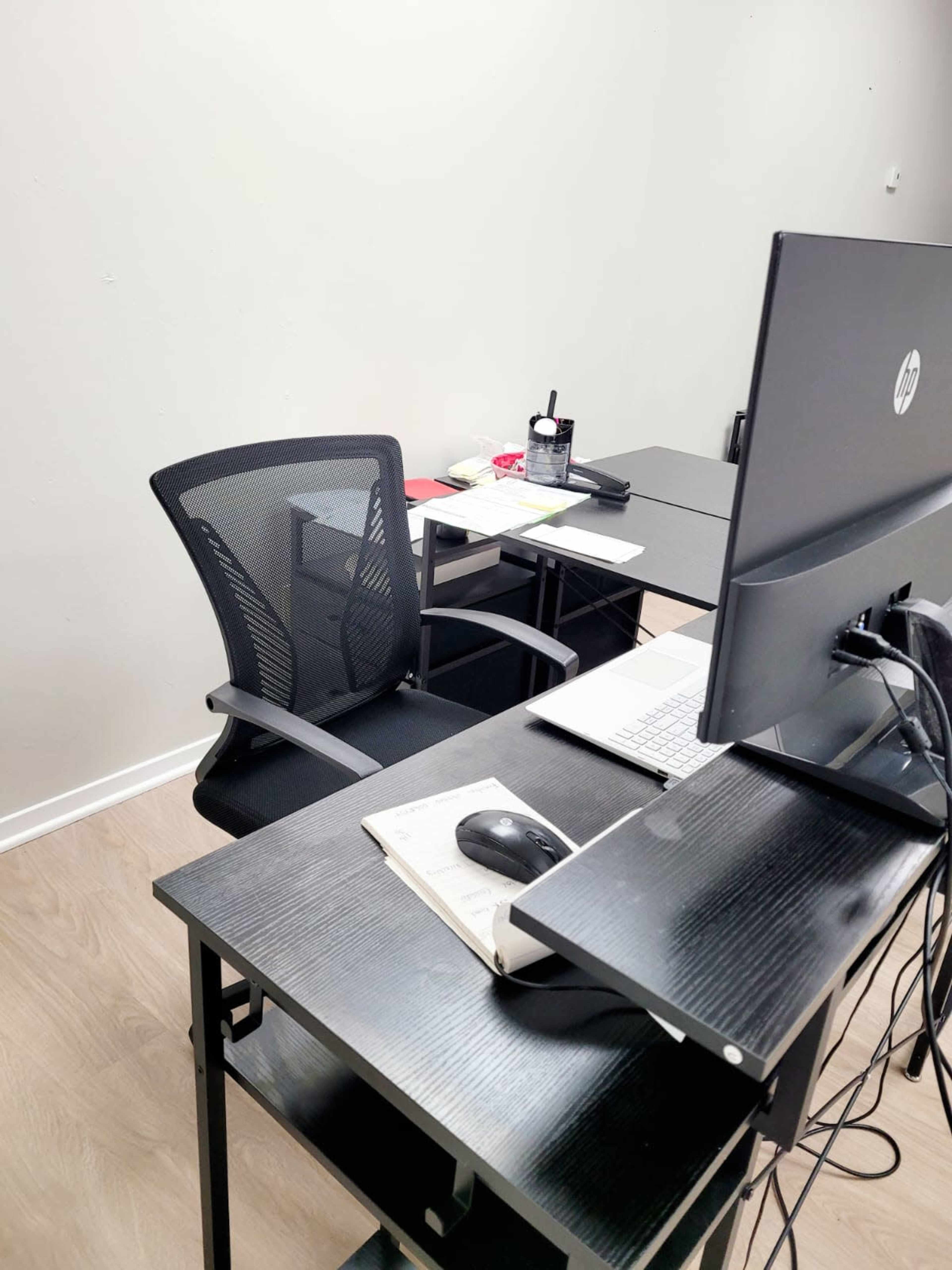 A black office chair is positioned in front of a computer monitor on a black desk, with a second desk nearby, both in a minimalistic workspace.