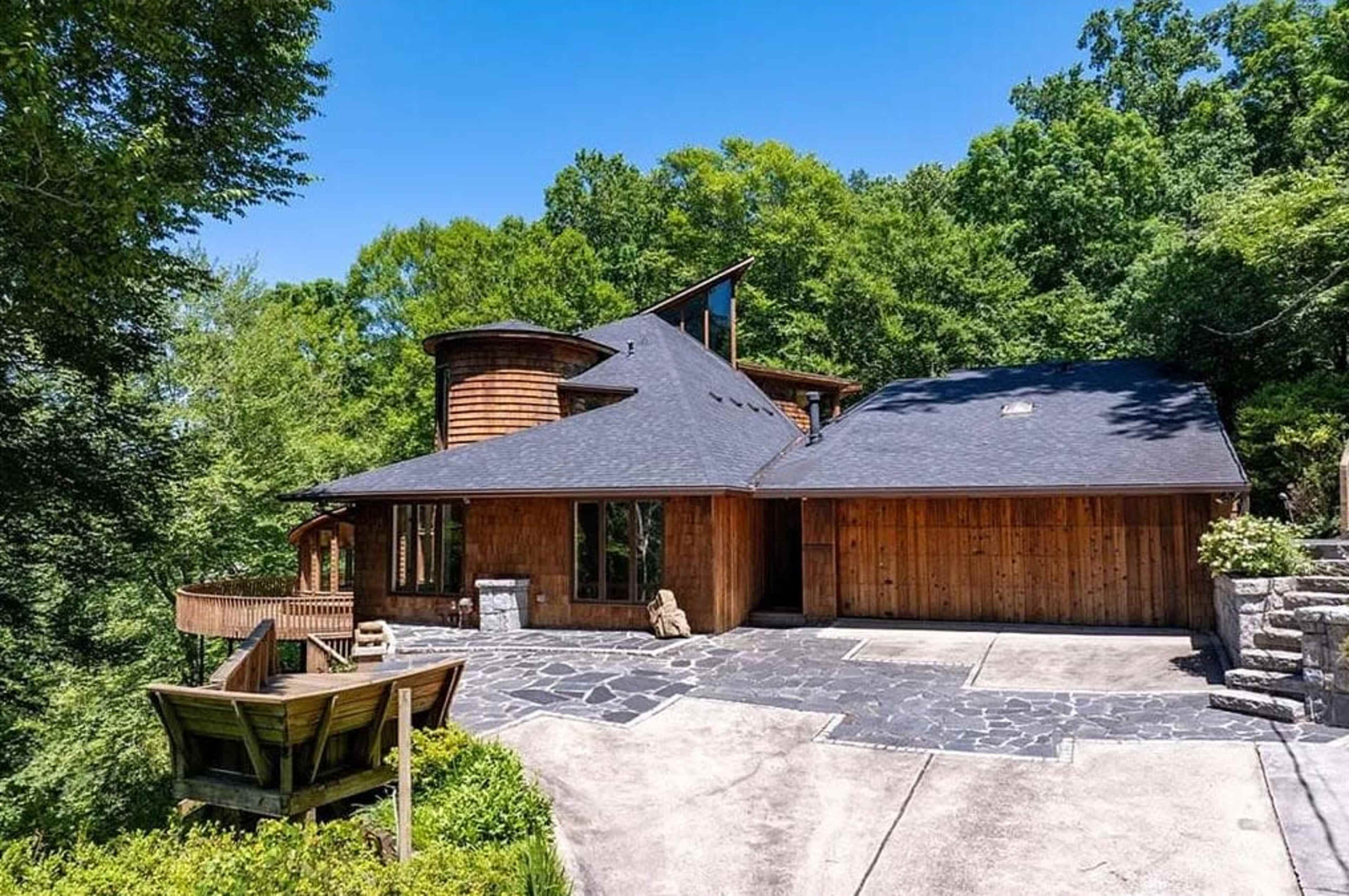 The image shows a modern, multi-level wooden house situated on a landscaped lot surrounded by trees.
