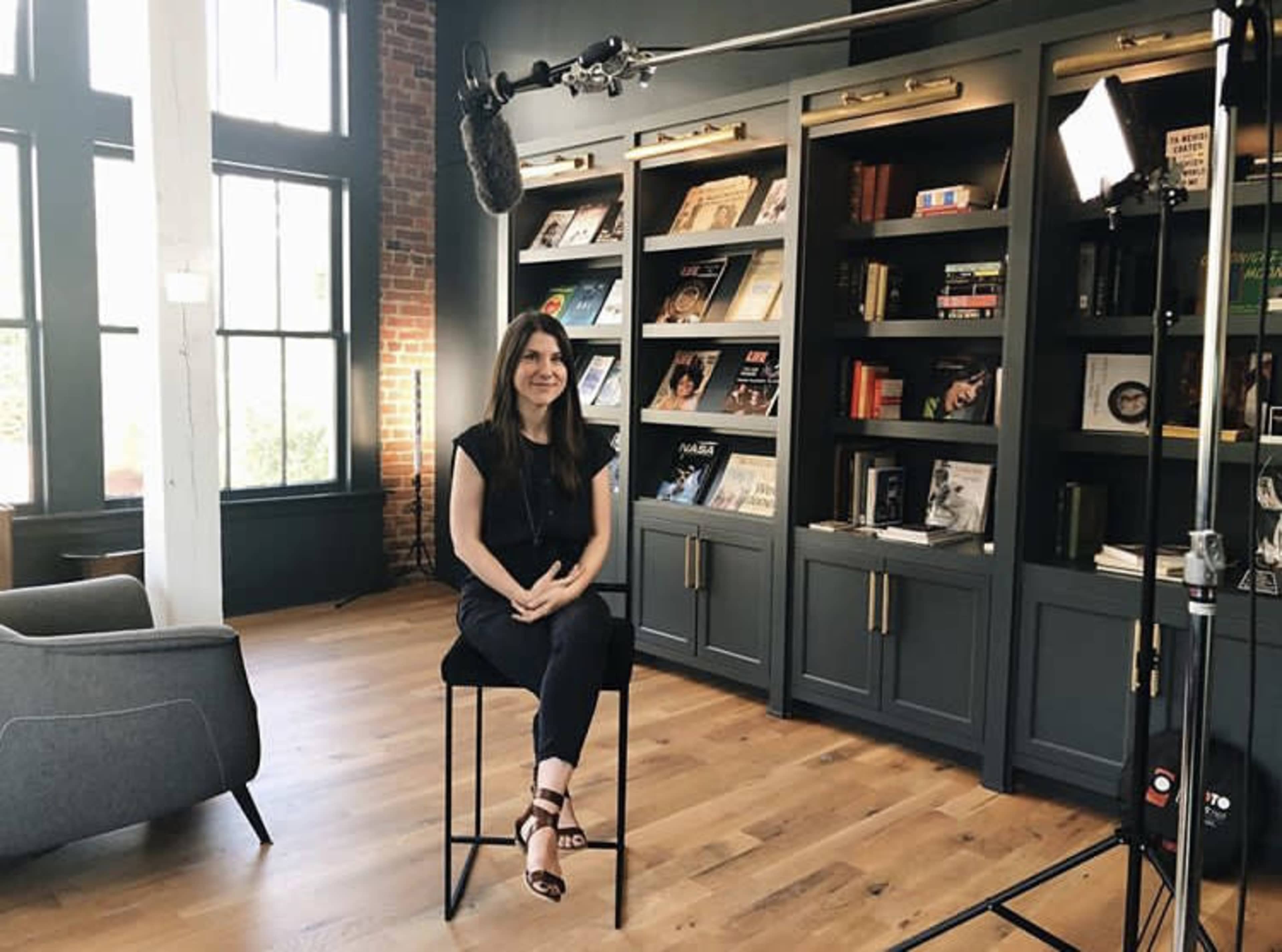 A woman sits on a bar stool in front of a bookshelf filled with books and a microphone is positioned above her in a well-lit room with large windows.