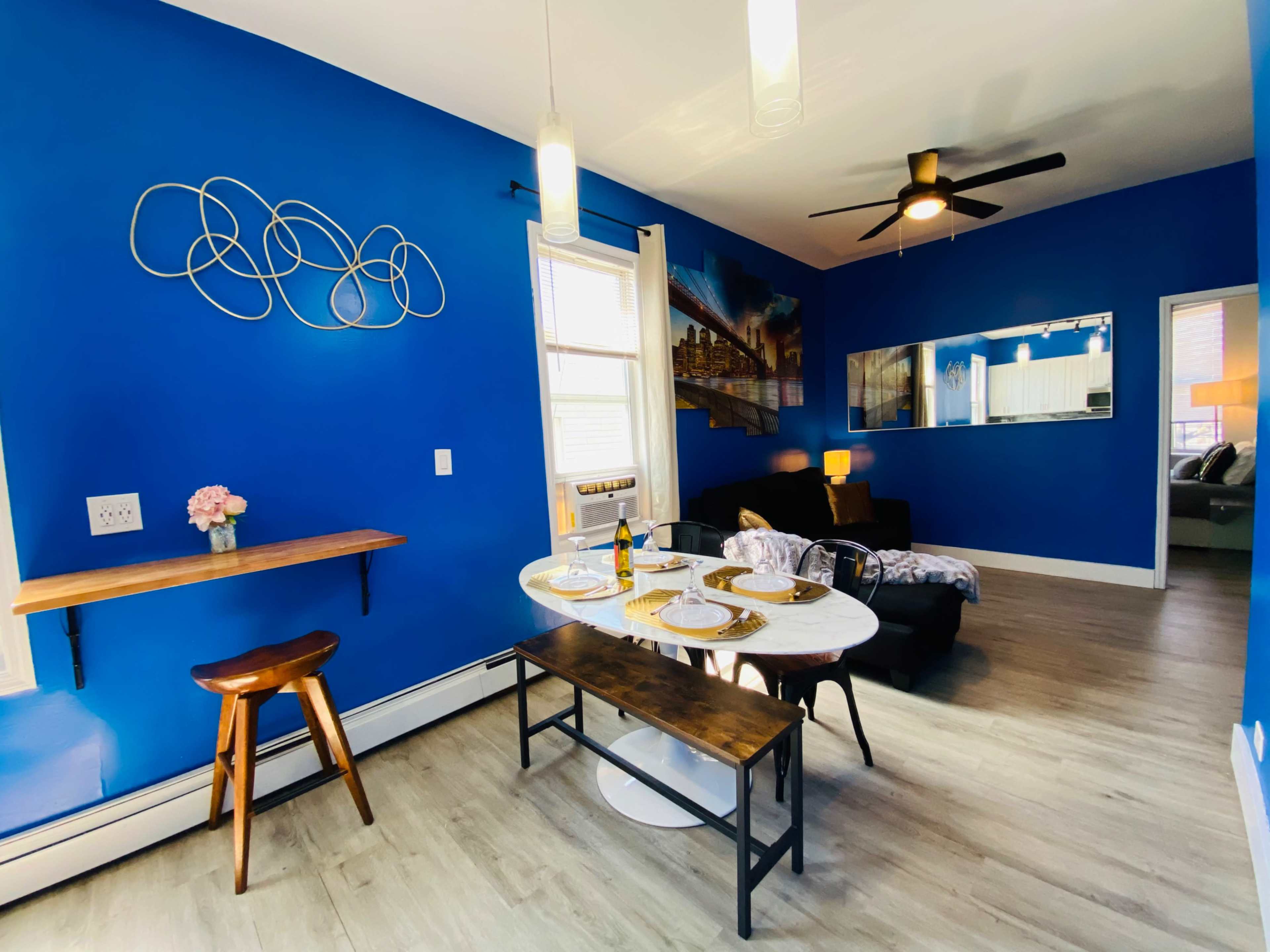 A dining area with a blue wall, a small table set for a meal, a wooden bench, and a living room visible in the background.