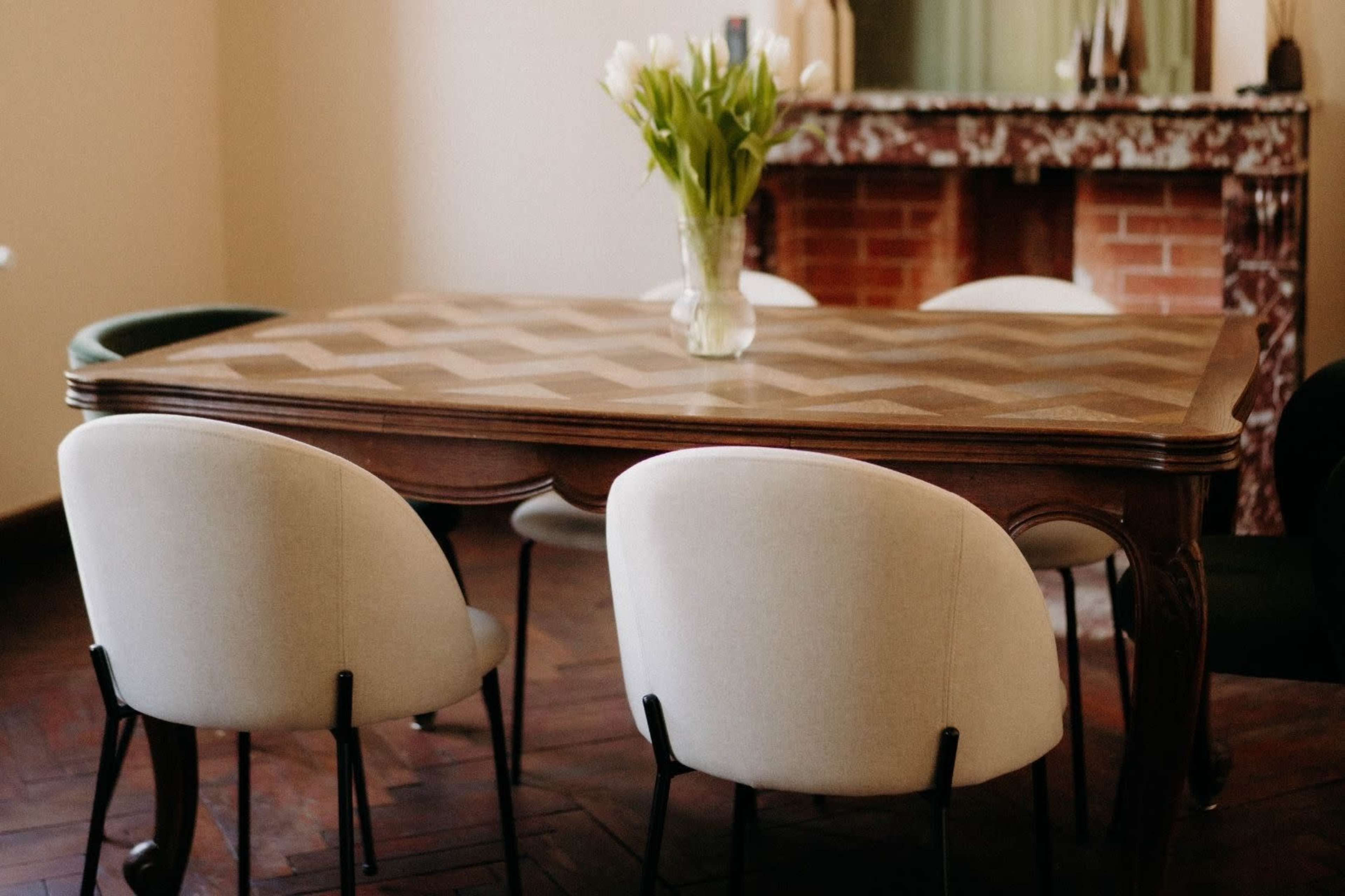 A wooden dining table with a chevron pattern is surrounded by upholstered chairs, while a vase of tulips sits in the center.