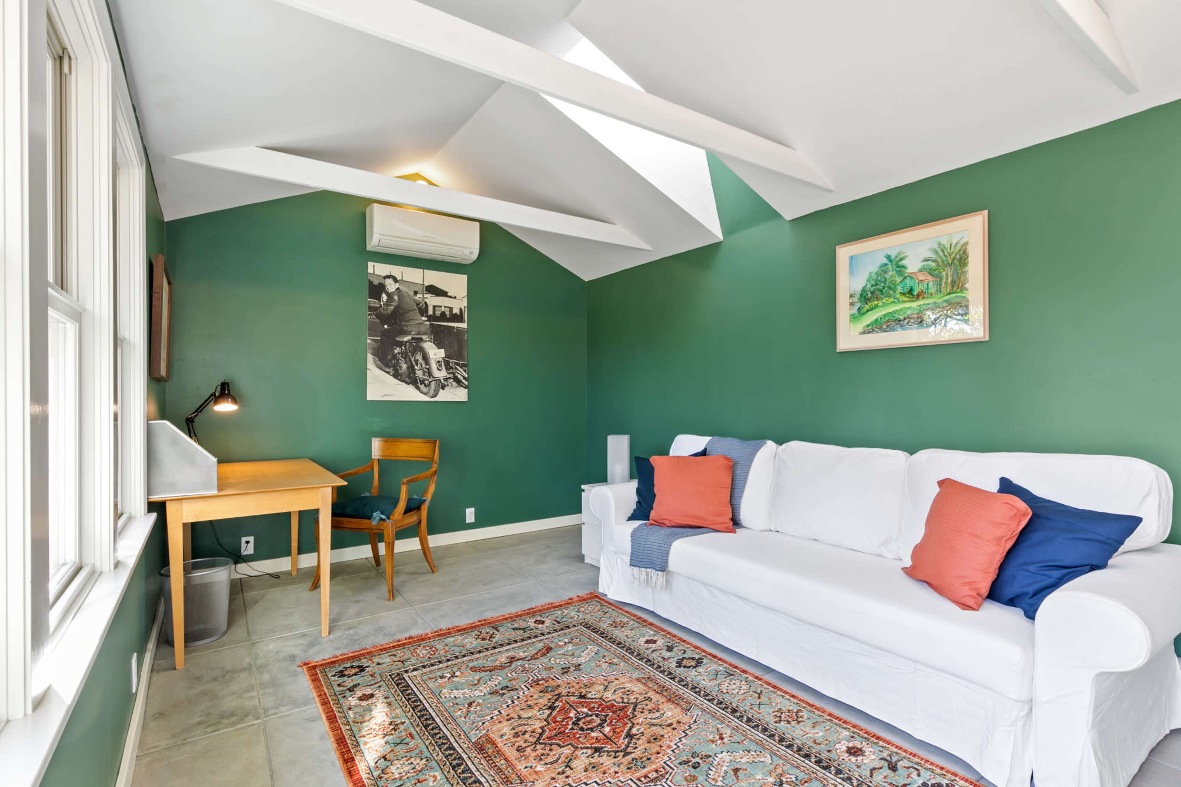 A bright room with green walls, a white sofa adorned with blue and orange cushions, a wooden desk and chair, and a patterned area rug on the tiled floor.