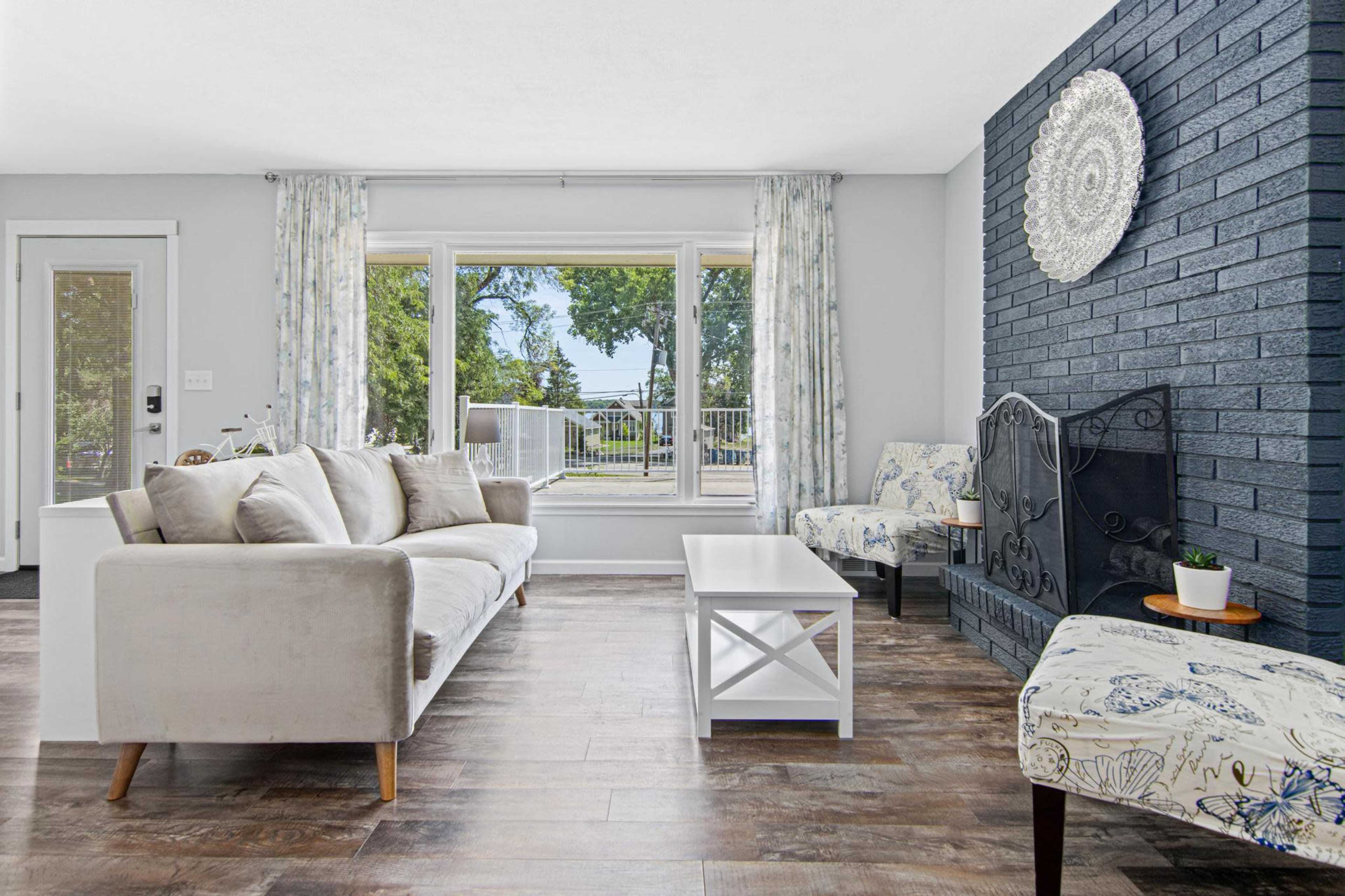 The living room features a light-colored sofa, a white coffee table, and two patterned chairs, with a fireplace and large windows overlooking a patio.