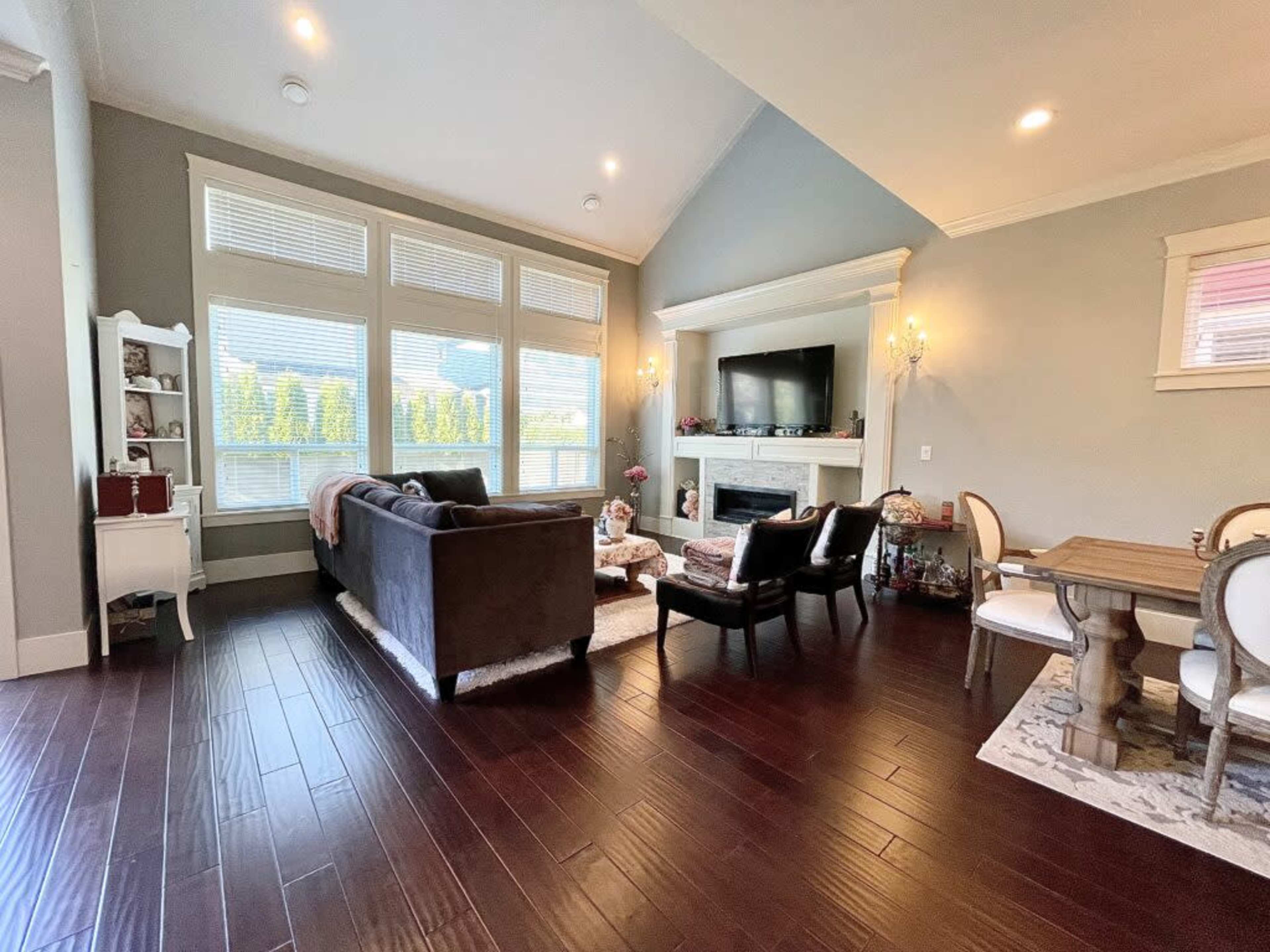 A well-lit living room featuring a dark sectional sofa, an armchair, a dining table, and a large television mounted above a fireplace, with large windows allowing natural light to fill the space.