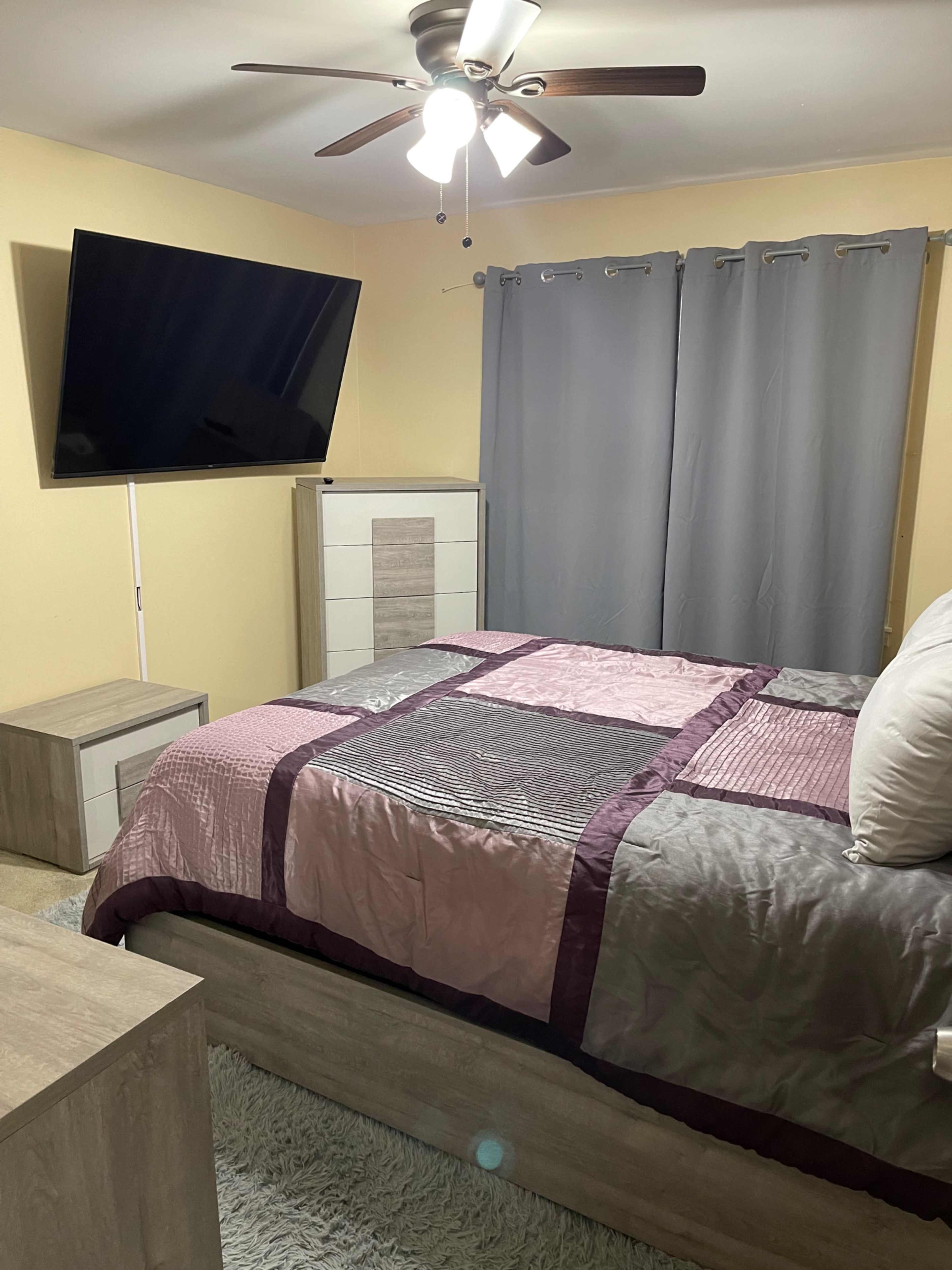 A bedroom with a large television mounted on the wall, a bed covered with a quilt, and gray curtains alongside a dresser and nightstand.