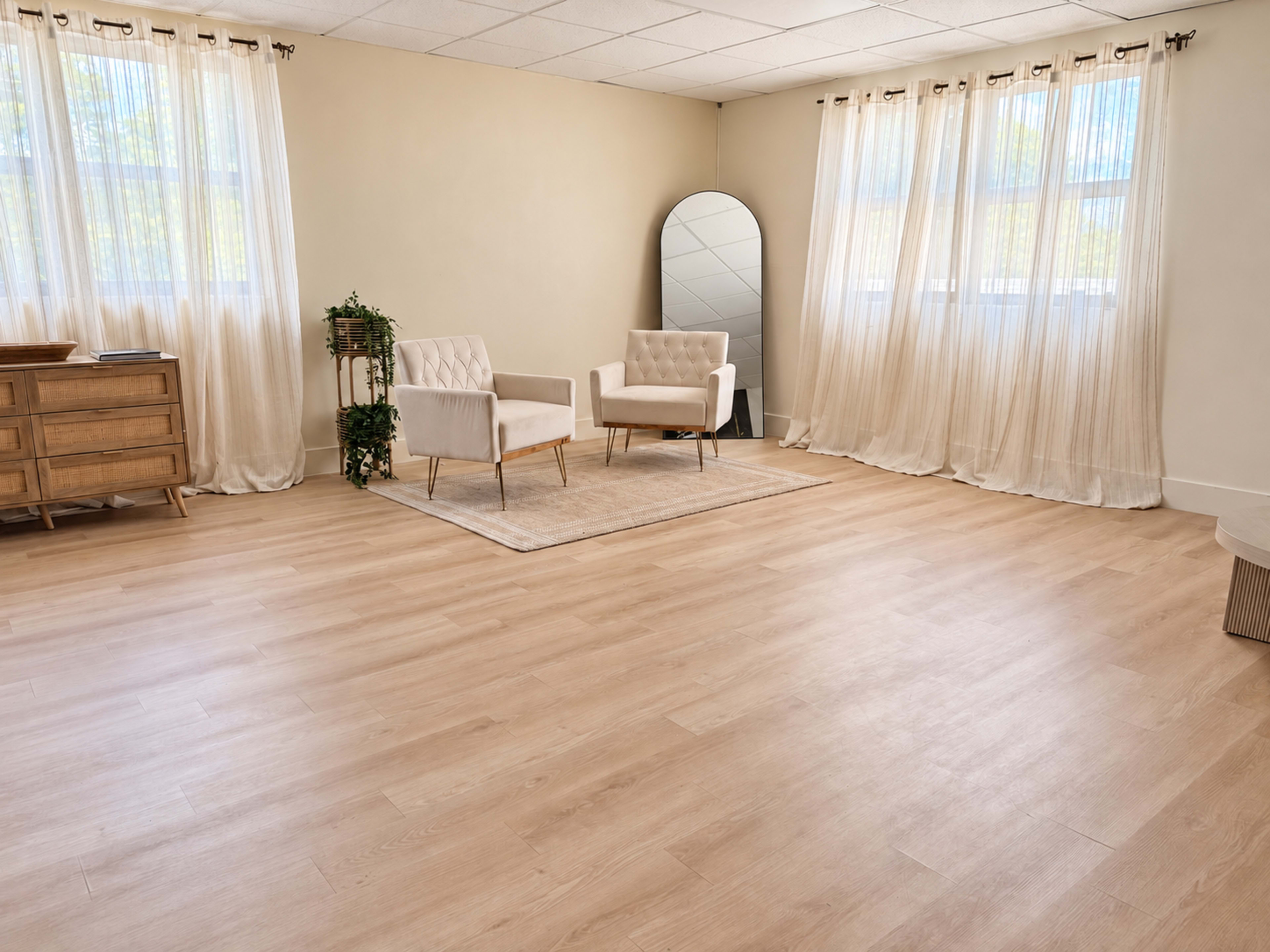 The image shows a spacious room with light wooden flooring, a large mirror, two upholstered chairs, and sheer curtains covering the windows.