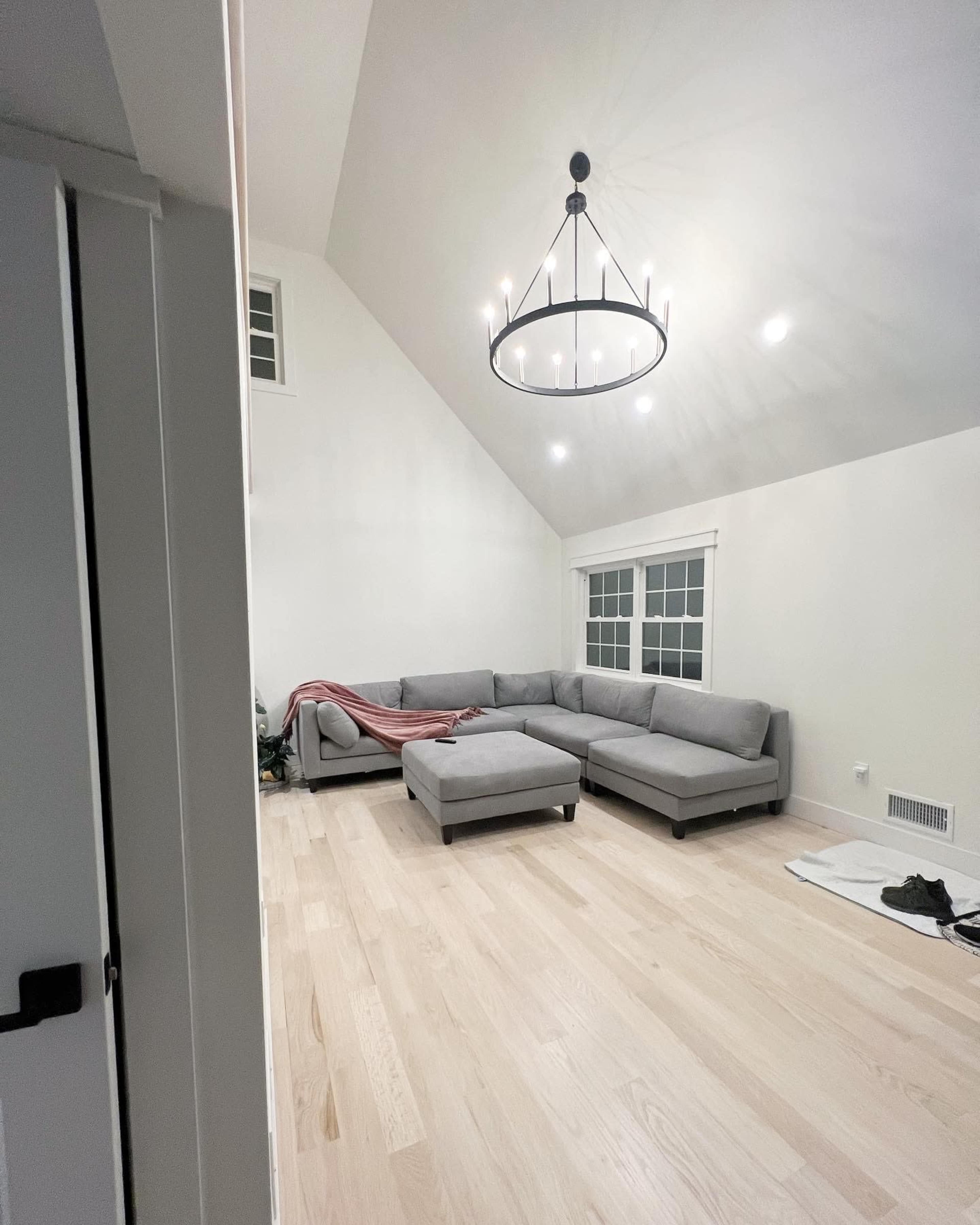 A spacious room with a high ceiling, featuring a gray sectional sofa, a round chandelier, and light hardwood flooring.