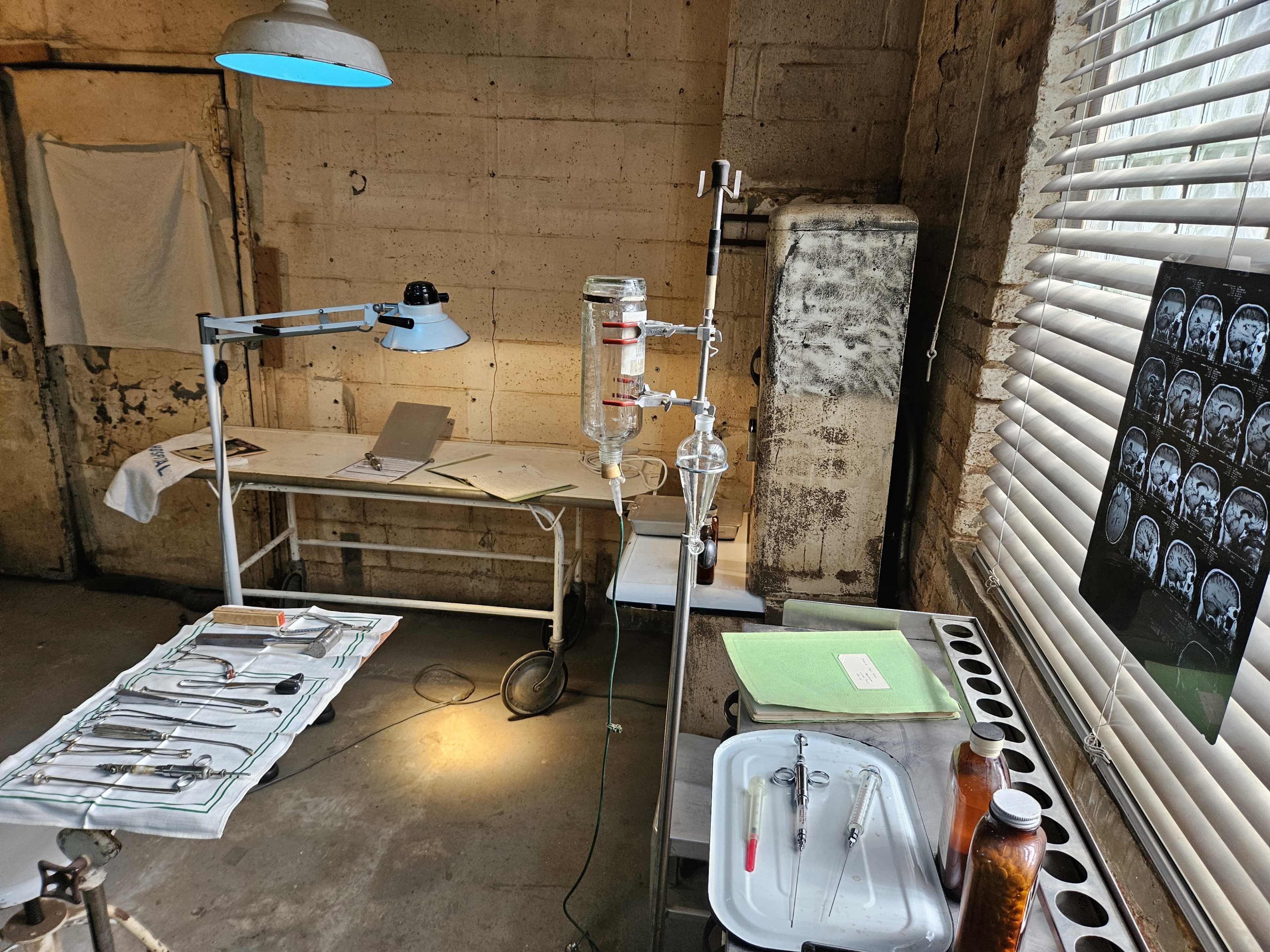The image depicts a medical examination room with surgical instruments arranged on a tray, a light fixture, and medical equipment, alongside a wall featuring X-ray images and a window with blinds.