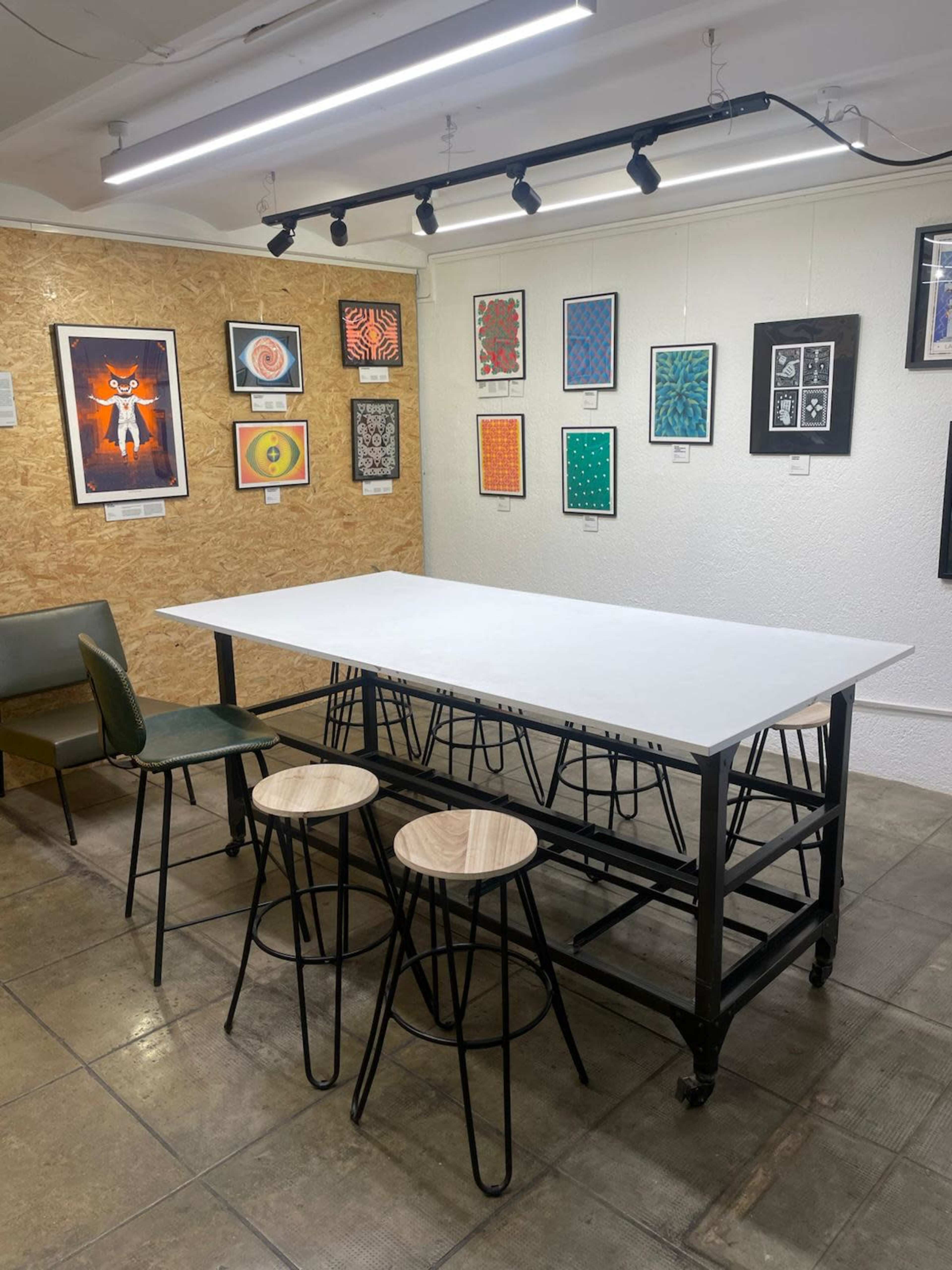 The image shows a simple art gallery featuring a large white table with four stools and multiple framed artworks displayed on the walls.