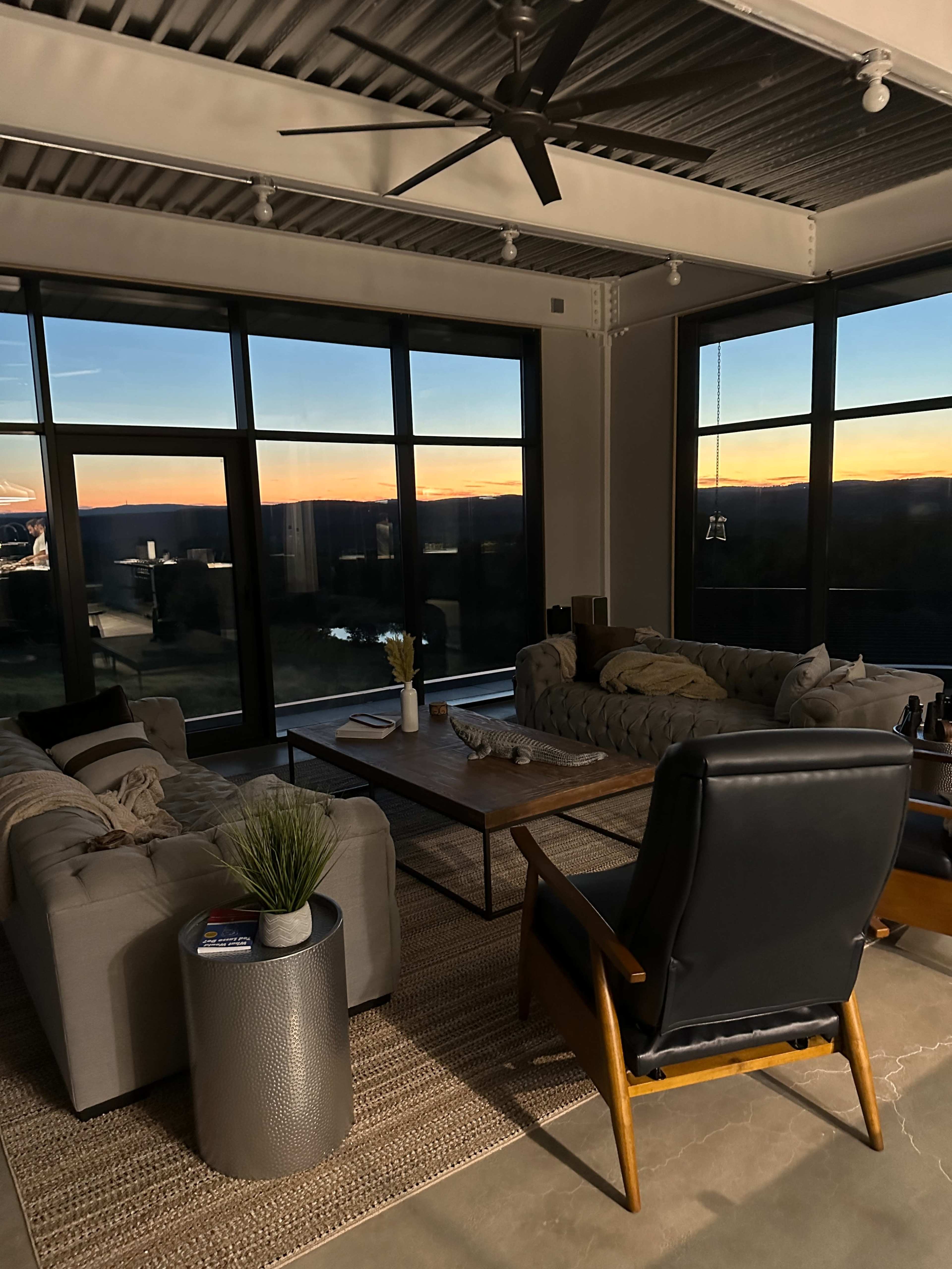 A modern living room with large windows overlooking a sunset, featuring a sectional sofa, a coffee table, and a black armchair.