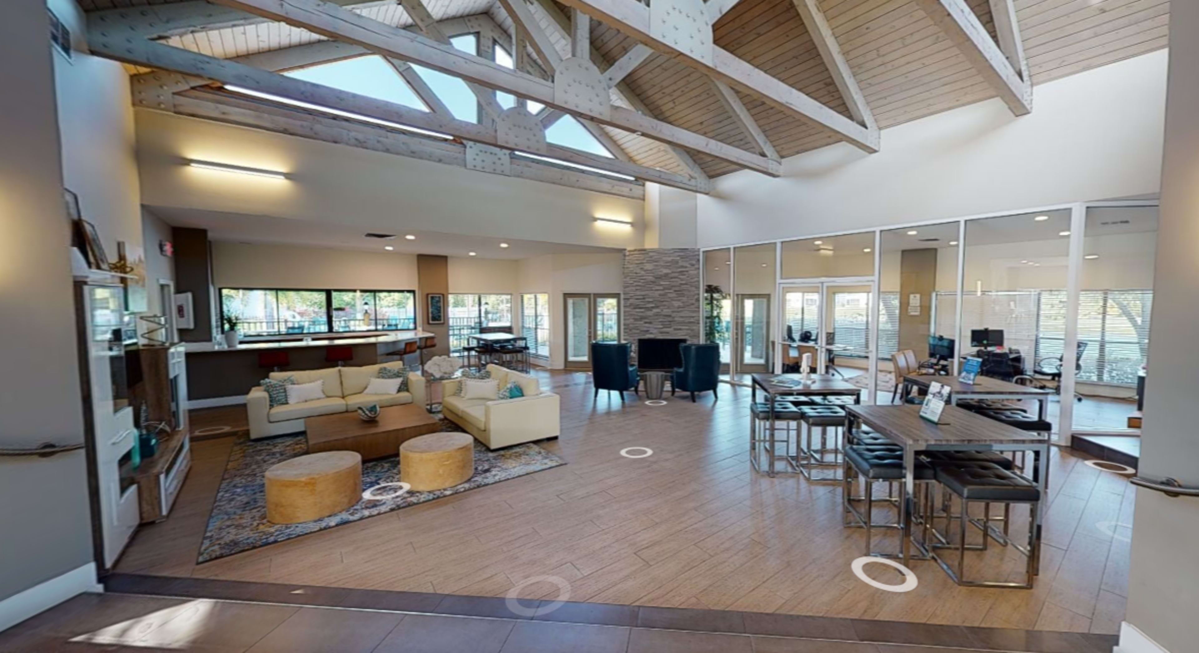 The image shows a modern community lounge with high ceilings, a seating area featuring circular ottomans, and a dining space with tables and chairs.