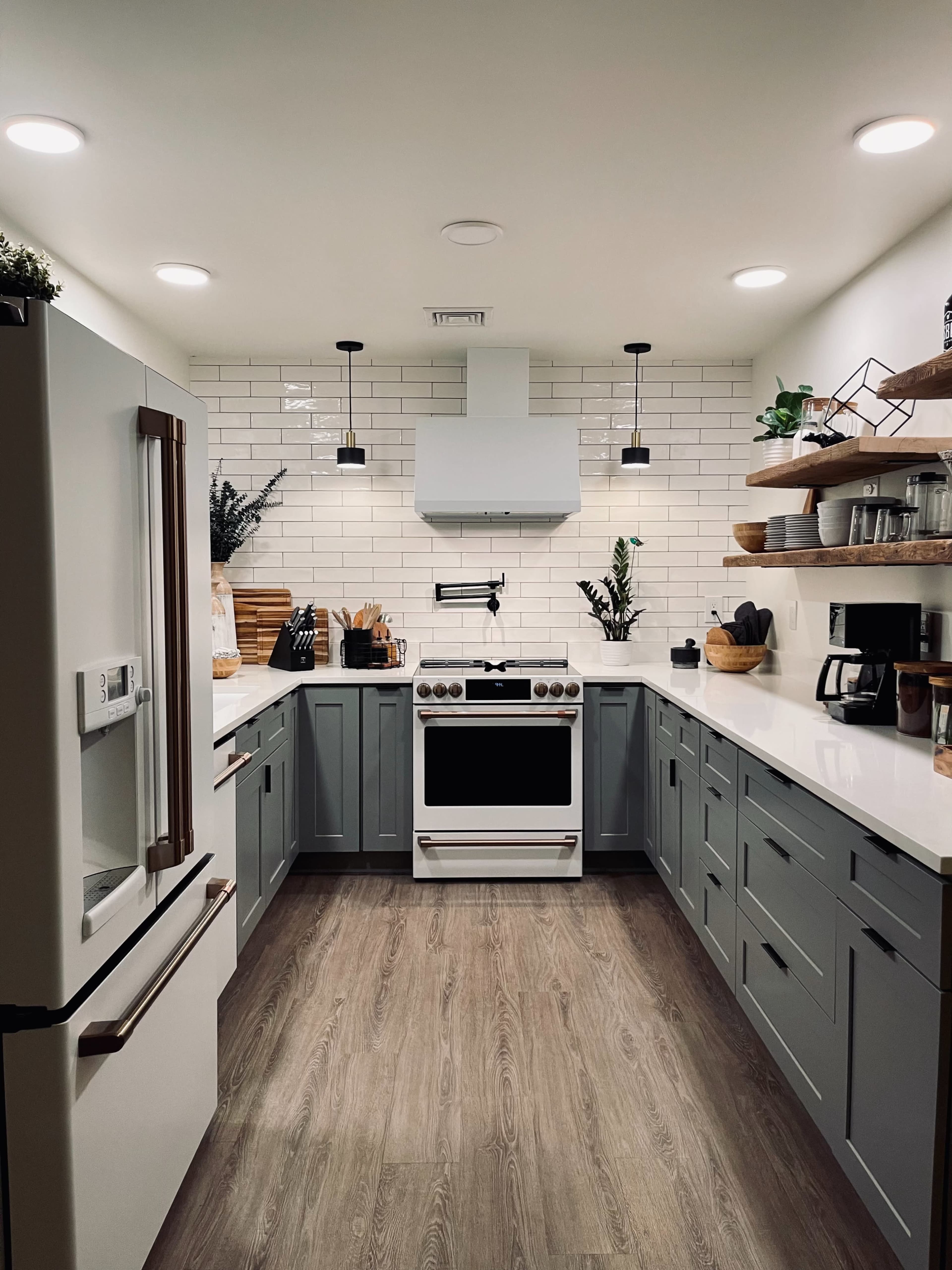 A modern kitchen features gray cabinets, a white stove, and a hood above a clean countertop with open shelving displaying kitchenware and plants.
