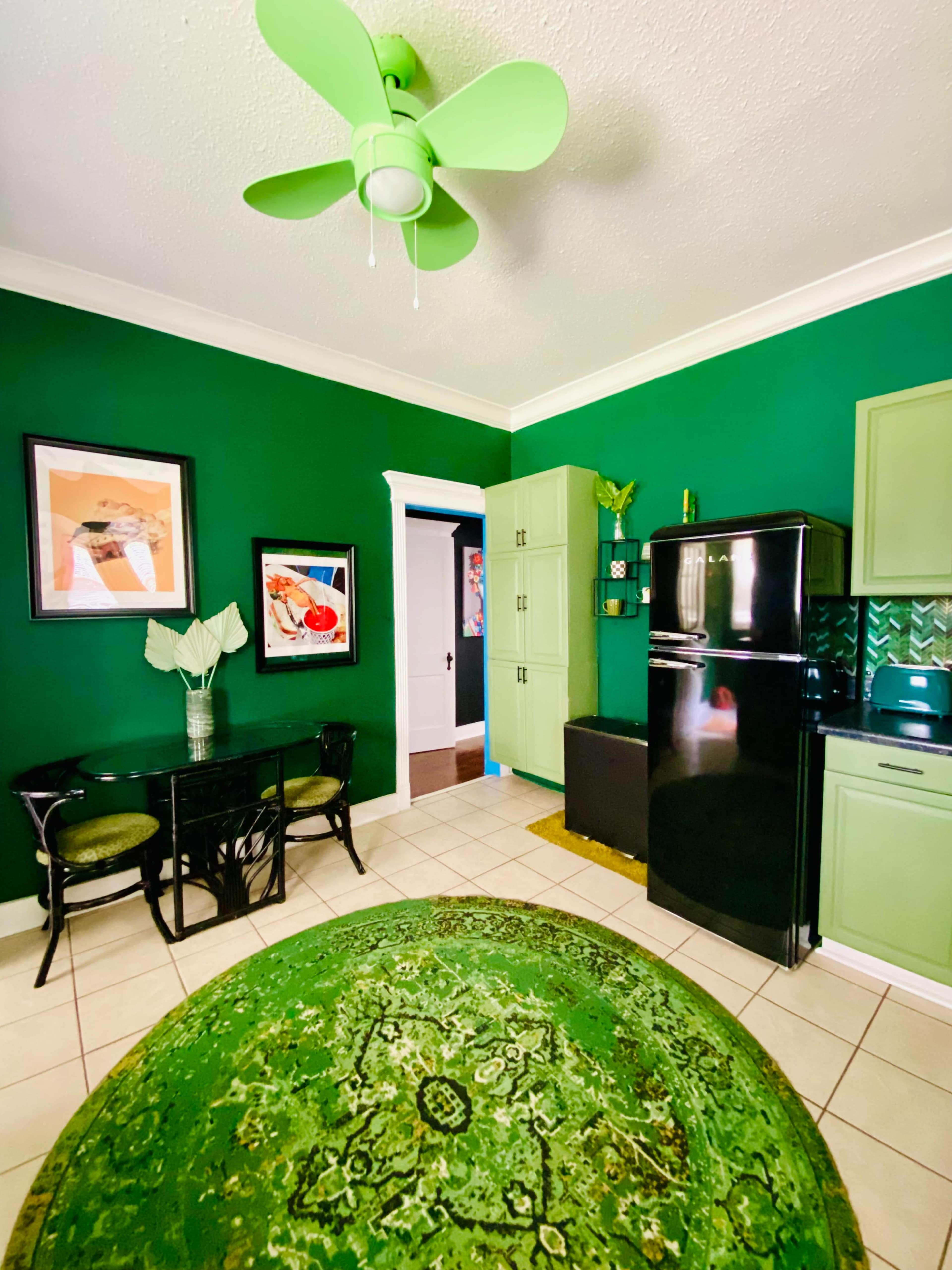A kitchen with green walls, a black refrigerator, green cabinetry, a round green rug, and a small dining table with matching chairs.