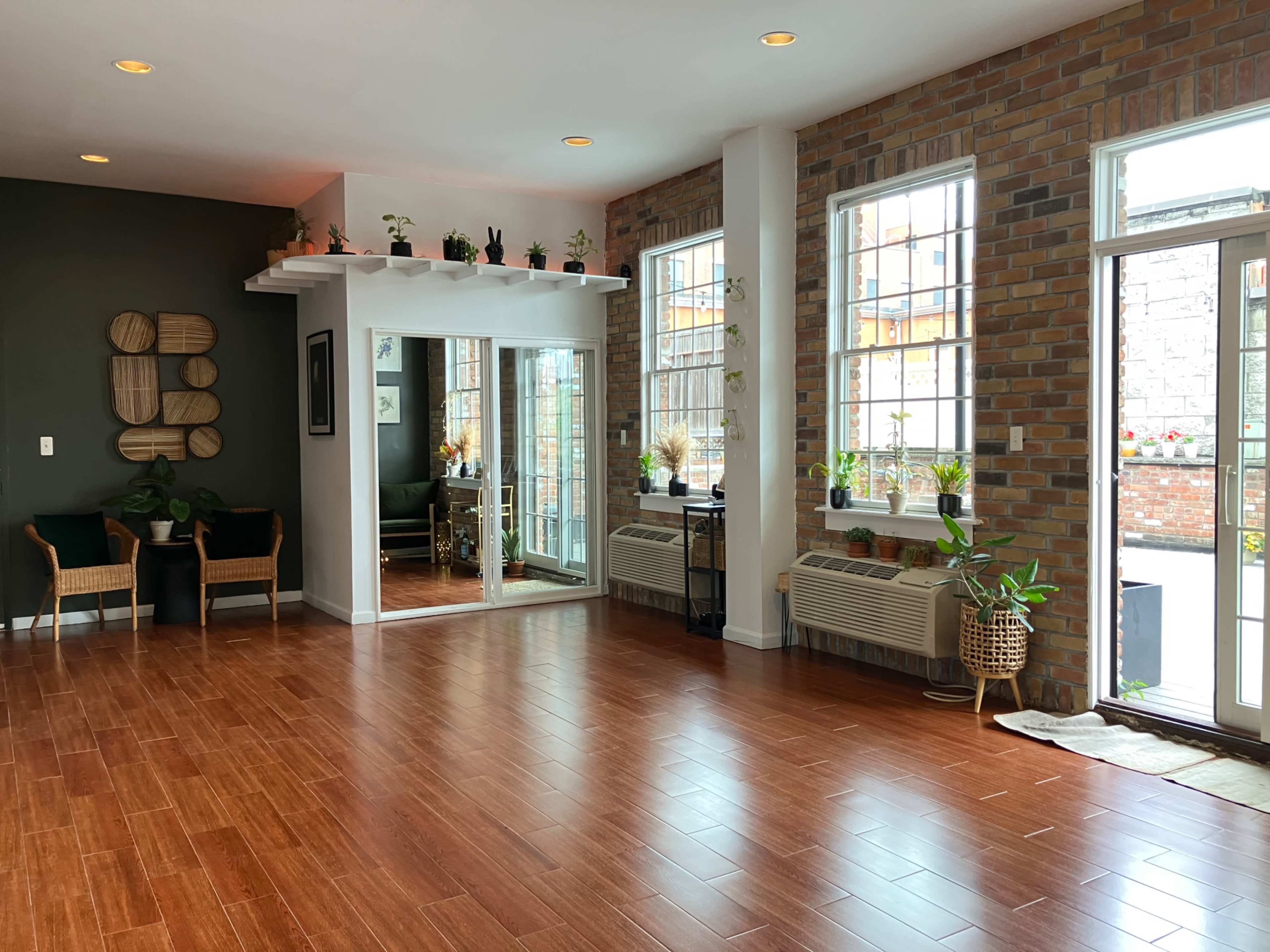 The room features hardwood floors, large windows with plants, and a mirrored wall reflecting a seating area.