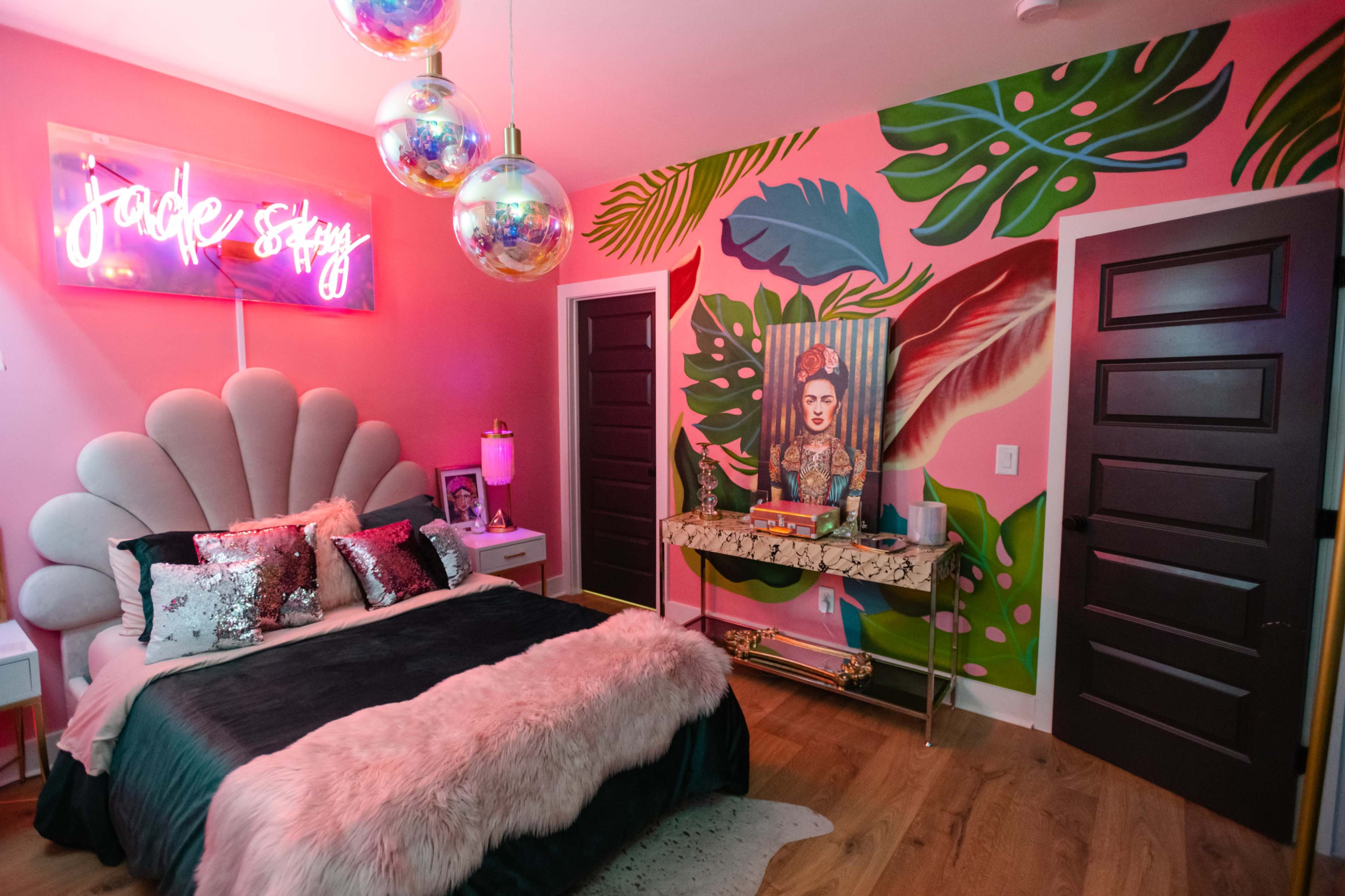 A brightly decorated bedroom featuring a large bed with a plush headboard, colorful wall art, and a neon sign that reads "jackie stacy."